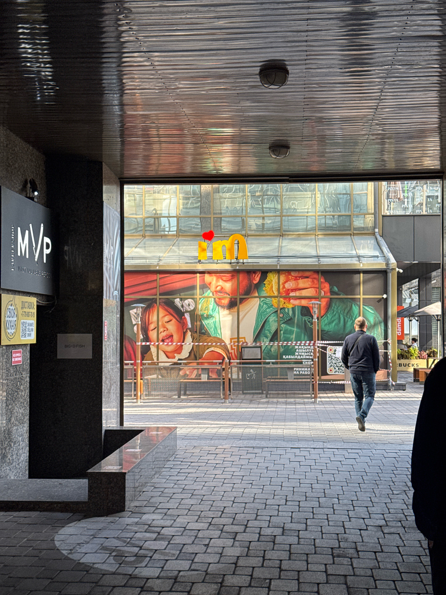 The image depicts an outdoor view of a pedestrian area, likely in Almaty, Kazakhstan, with a large advertising mural on a building facade. The mural features a close-up of a man holding a large hamburger, with a young girl to his left. The mural is framed by large windows. Above the mural, yellow lettering with a red heart is visible. In the foreground, a man wearing a dark jacket and blue jeans walks away from the camera. The ground is paved with grey cobblestones. To the left of the frame, a portion of a building with signage is visible, including a black sign with "MVP" in white lettering, and below it, yellow signs with Cyrillic text. A reflective ceiling made of corrugated metal spans the top of the frame, with two light fixtures visible. In the background, there are tables and chairs, suggesting an outdoor seating area for a cafe or restaurant. The overall scene appears to be a public walkway leading to an establishment, possibly a fast-food restaurant given the imagery on the mural.