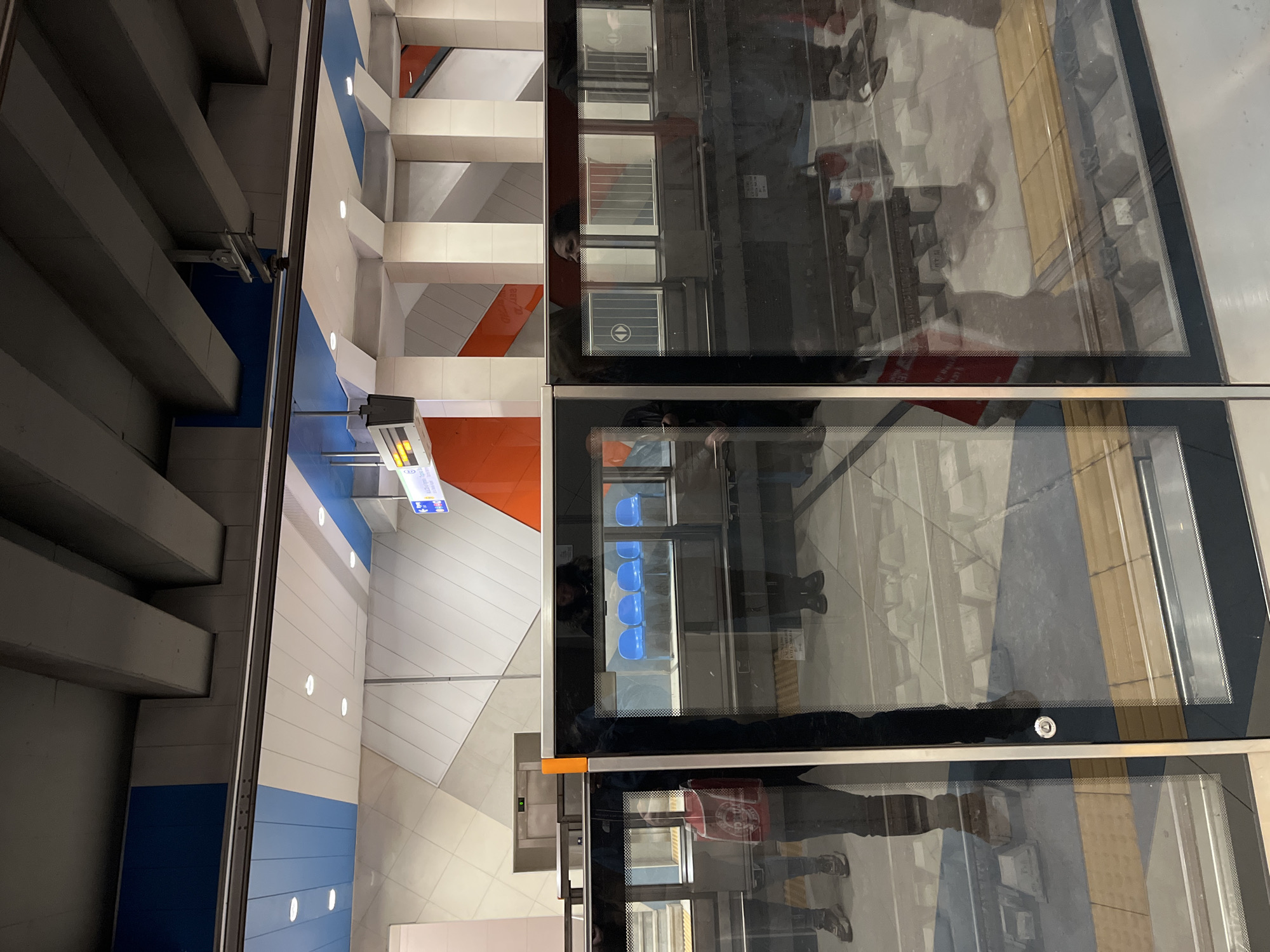 The image depicts an elevated view of a subway station in Sofia, Bulgaria. The station's architecture features a modern design with blue, white, and orange color schemes on the walls and ceiling. The platform is visible through glass barriers, reflecting the activity on the platform, including individuals, seating, and signage. The top half shows the ceiling and architecture, while the bottom half shows the platform.