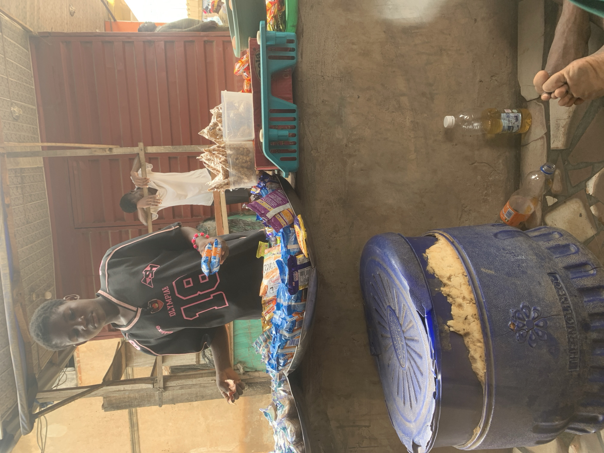 In Shagamu, Nigeria, a young person stands behind a market stall, holding packets of snacks. The stall features a variety of packaged biscuits and crackers displayed on trays. In the background, another person leans on a wooden structure, and a third person stands further back. A large blue insulated container is visible in the foreground, and a bottle of liquid and some empty plastic bottles are on the ground. The environment appears to be an outdoor market setting with a corrugated metal structure as part of the stall.