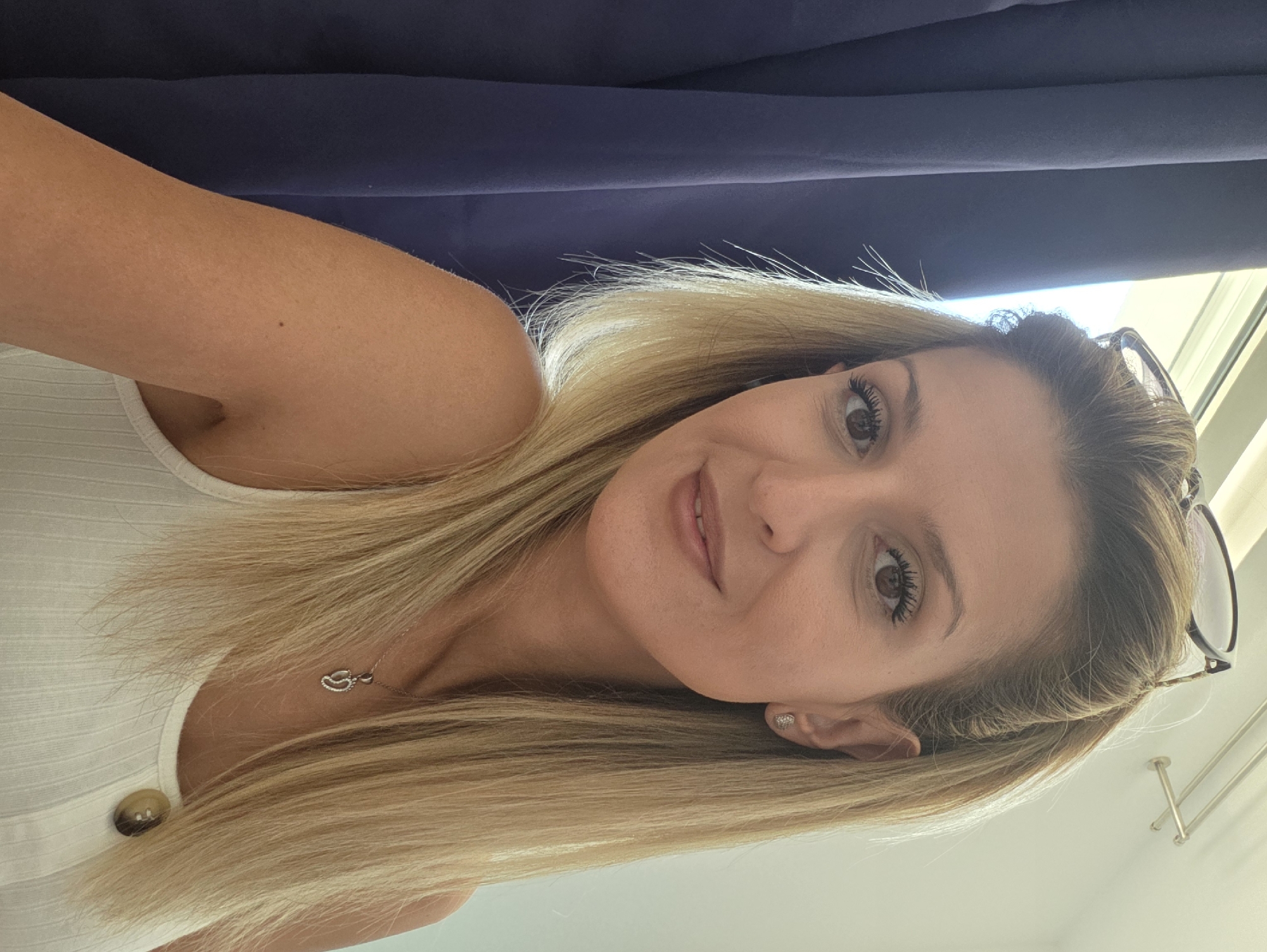 A woman with blonde hair and brown eyes is looking at the camera. She is wearing a white tank top with a button and a silver necklace with a pendant. She has on a pair of glasses on top of her head. She is standing in front of a dark blue curtain. The background appears to be a room with a window and a light-colored wall. There is no indication of the specific location within Munich, Germany.