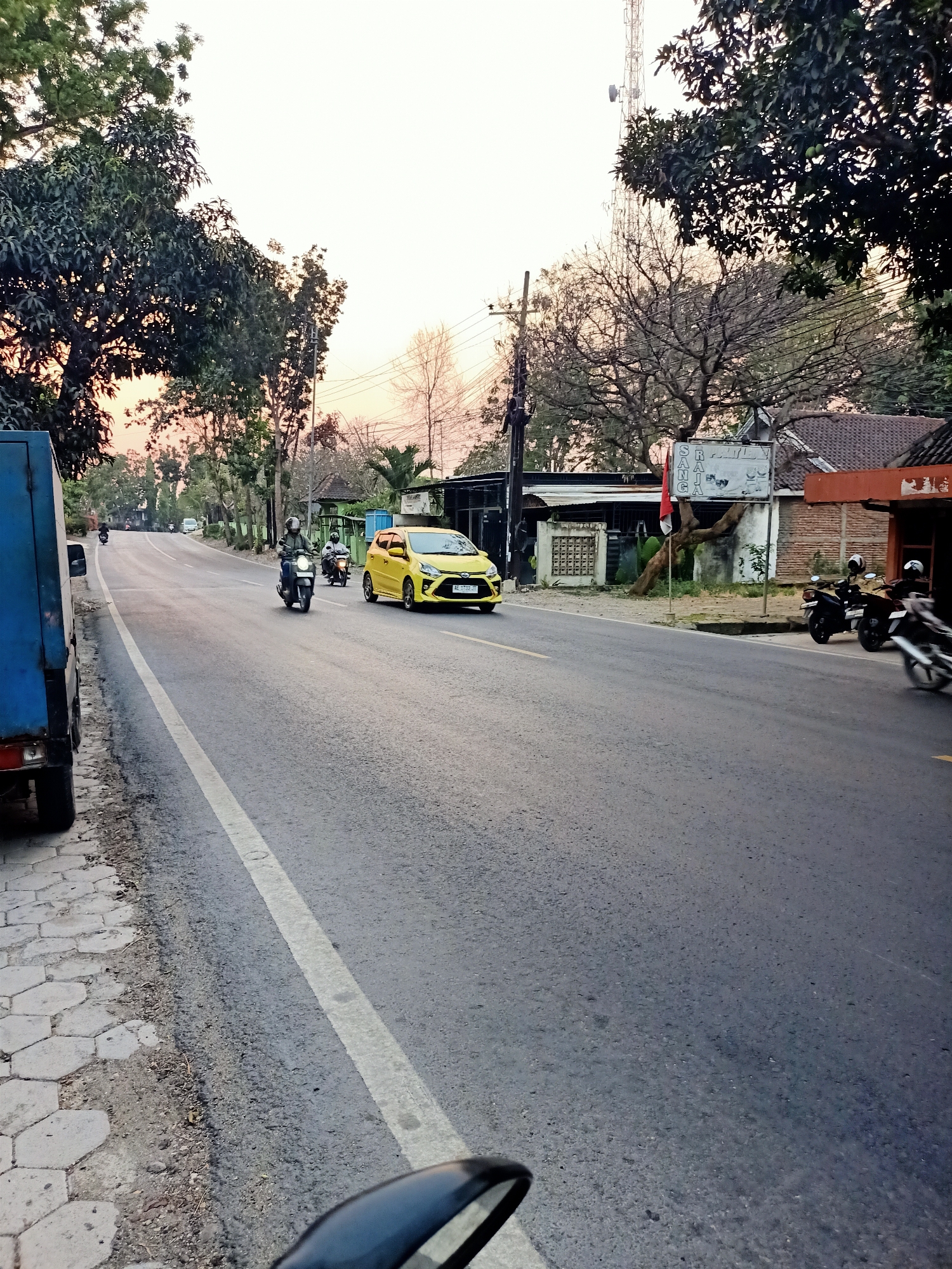The image depicts a street scene in Kendung, Indonesia, likely during the late afternoon or early evening given the soft light and warm tones in the sky. The road is paved and has a white dividing line. Several motorcycles are visible on the road, some in motion and others parked. One motorcycle in the foreground is moving towards the right, with a rider wearing a helmet. Further down the road, a yellow car is also in motion. A blue truck is parked on the left side of the road, partially in frame. The street is lined with trees on both sides, some with dense foliage and others appearing bare. Buildings with tiled roofs are visible along the right side of the road, with a red and white flag planted in front of one. Utility poles and wires are also present. The overall environment suggests a suburban or semi-rural setting within the city.