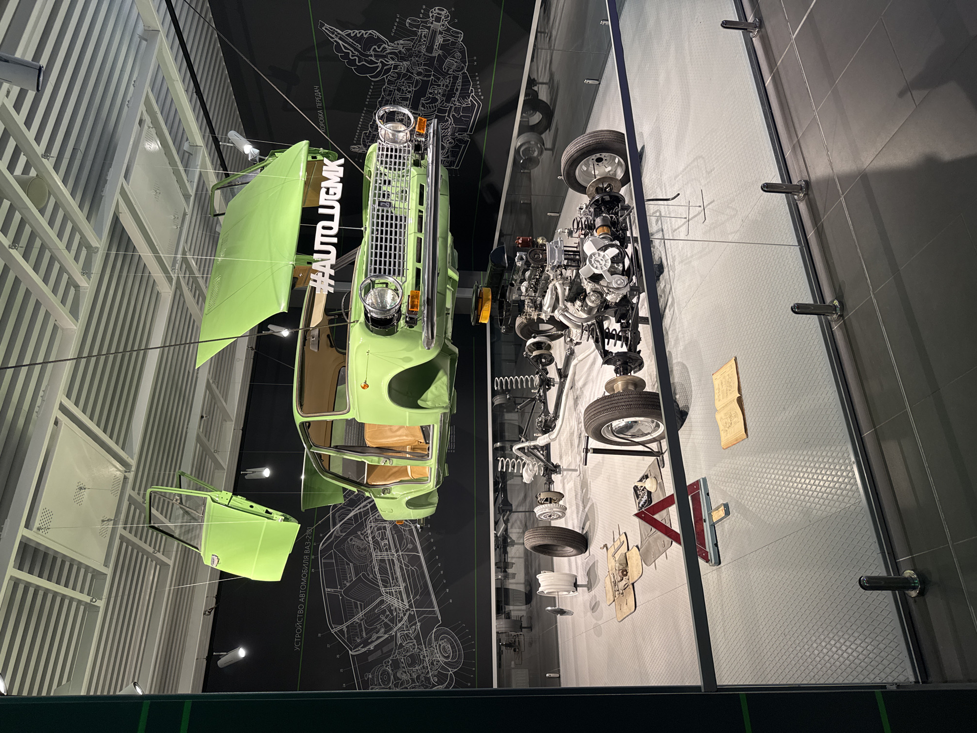 The image captures an exhibit at the UMMC Museum Complex in Verkhnyaya Pyshma, Russia. A disassembled light green vehicle is suspended from the ceiling. The parts include the vehicle's body, hood, door, chassis, engine, wheels, and other components. The vehicle is suspended over two adjacent wall sections. One wall has a black background with a blueprint-style technical diagram of the vehicle. The adjacent wall is covered in light grey tiles. Mechanical parts from the vehicle are affixed to the light grey tiles. A book, road hazard triangle, and other loose parts are also affixed to the wall. The ceiling above the left side of the vehicle features white horizontal lines. A section of the green framework is visible in the foreground.