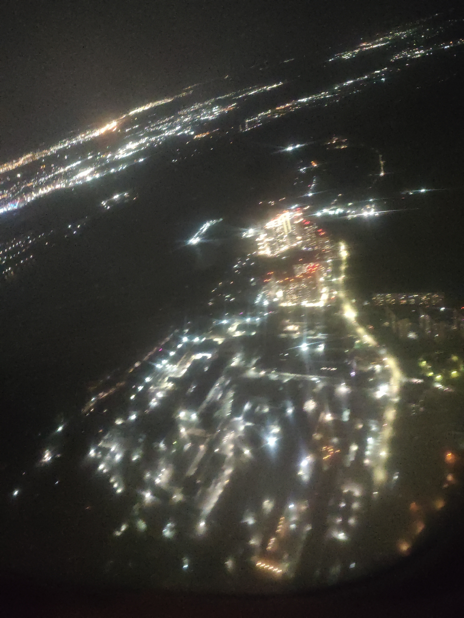 The image shows a nighttime view of a city, likely Novosibirsk, Russia, as seen from an elevated position, possibly an airplane. The city is illuminated with various lights, creating a network of brightness against the dark background. The lights appear to be from buildings, streets, and other urban infrastructure. The scene is somewhat blurred, indicating movement during the photograph's capture.