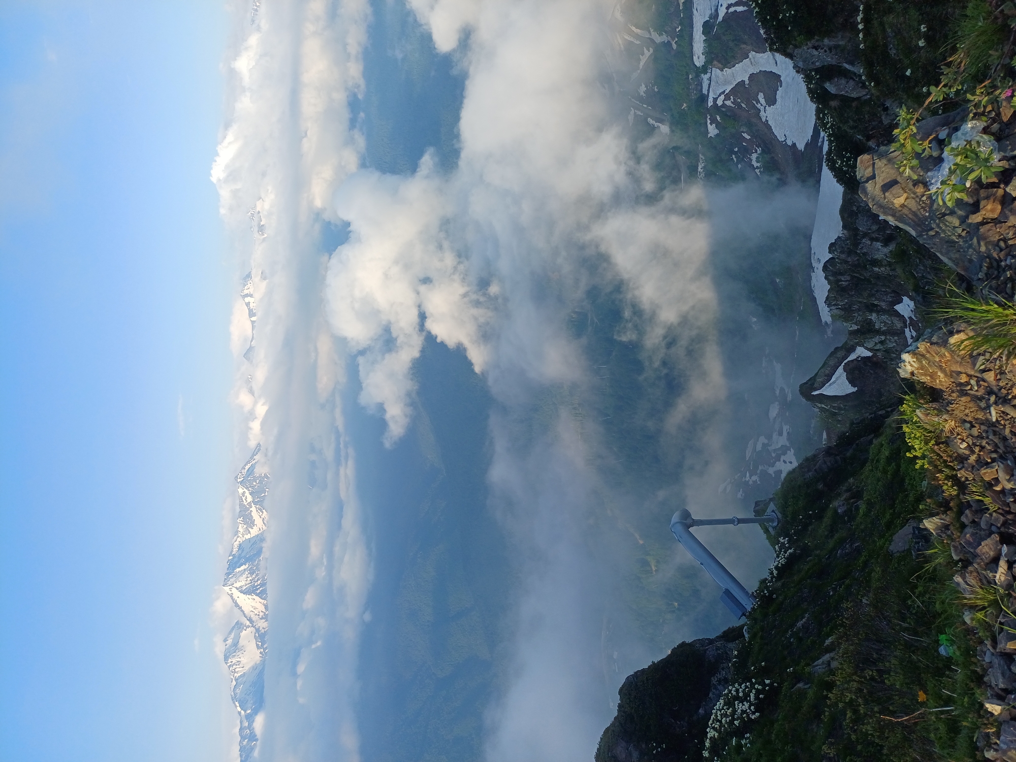 A high-angle shot captures a majestic mountain landscape. In the foreground, a rocky, overgrown slope descends steeply. A prominent, grey pipe system, supported by a metal structure, is visible on the cliffside, with solar panels attached. White wildflowers dot the green foliage on the left and right sides of the frame. The middle ground features a valley partially obscured by swirling white clouds and mist, with hints of green forestation. In the distance, a range of snow-capped mountains pierces the clear blue sky. The overall impression is one of rugged natural beauty and a touch of human engineering against a vast mountainous backdrop. This scene is likely located in or around Estosadok, Russia, a popular mountain resort area.