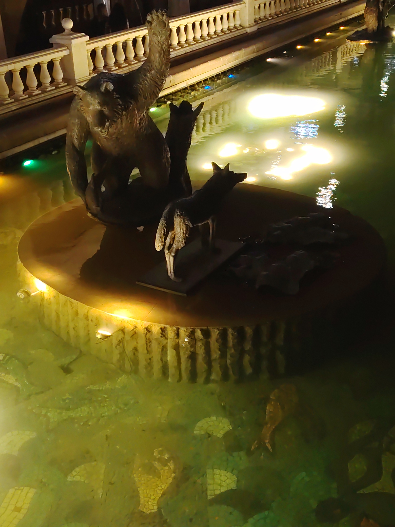 A bronze sculpture of a bear is positioned in a fountain, with its left arm raised. To the bear's right, a smaller bronze sculpture of a wolf stands on a circular stone platform that is partially submerged in the fountain's water. The fountain's water is illuminated by artificial lights, creating glowing reflections on the surface. In the background, there is a white balustrade and a building facade. The bottom of the fountain is decorated with mosaics.