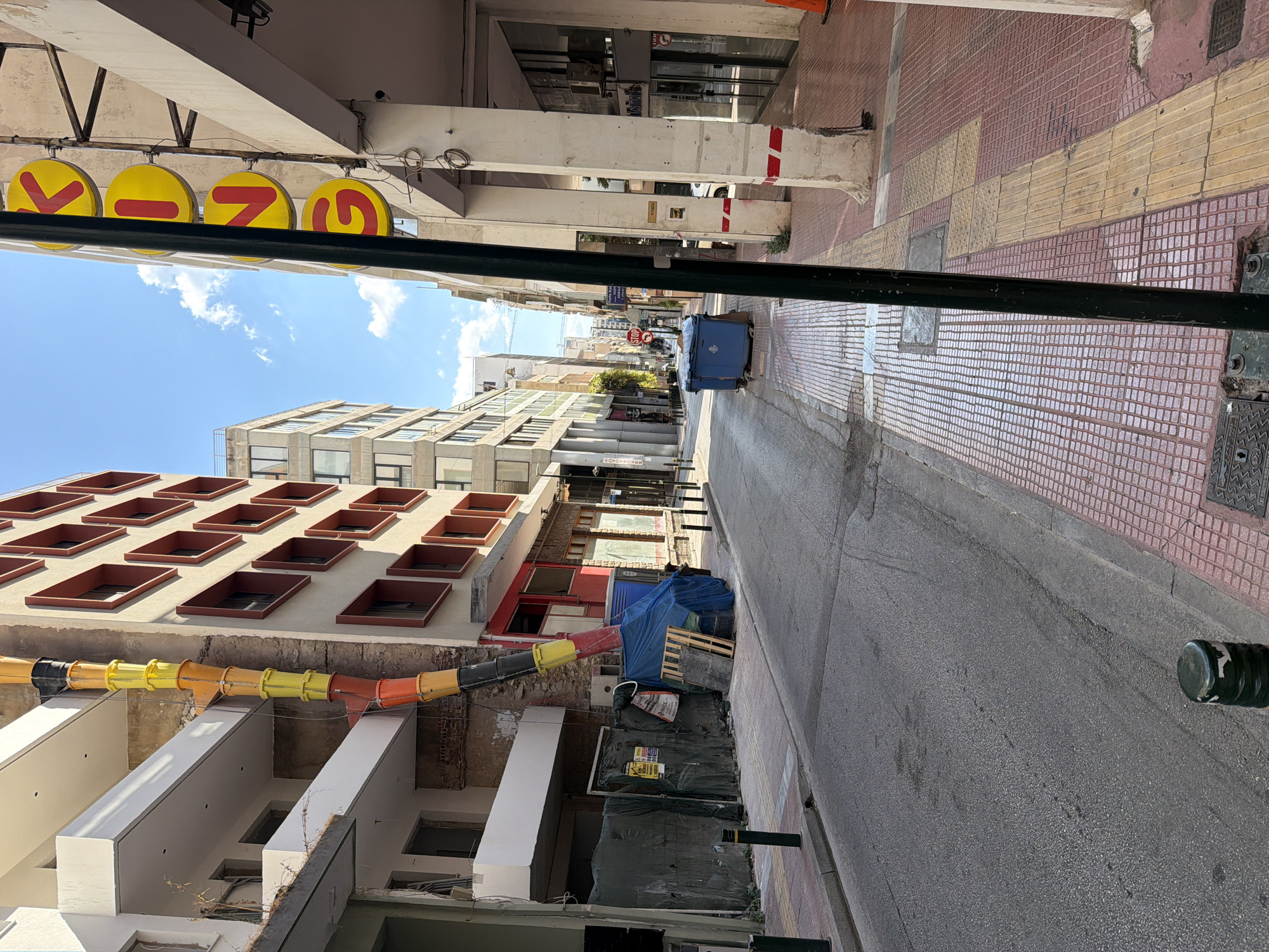 The image depicts a street scene in Piraeus, Greece. A narrow street is visible, lined with buildings on both sides. The left side of the street features a building with balconies and exposed brickwork, with a colorful debris chute extending downwards from an upper level. Construction materials, including pallets and a blue tarp covering some items, are present along the sidewalk.
The right side of the street has a colonnade with large white pillars. Above the colonnade, a sign with large yellow circular letters spelling "KING" in red is mounted vertically. The sidewalk on the right is paved with red and white tiles, with a section of textured yellow tiles forming a tactile paving strip.
Further down the street, two large blue dumpsters are situated on the right side. The street itself is asphalt and appears to be in a residential or commercial area. The sky is blue with scattered white clouds. A black pole runs vertically in the foreground, obscuring a portion of the view. The overall impression is that of an urban environment, possibly undergoing some form of renovation or construction.