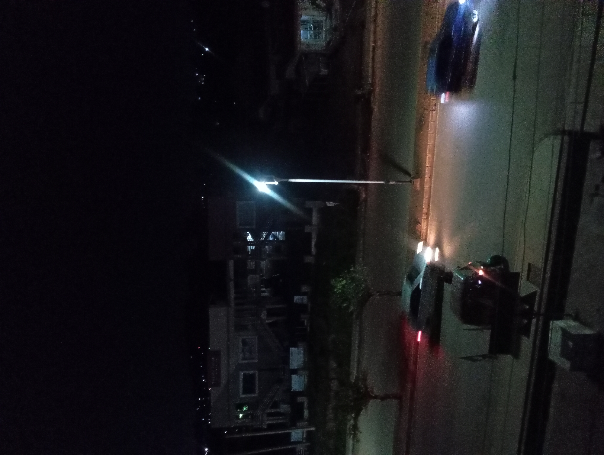 An outdoor, high-angle, nighttime photograph shows a street scene in an urban environment. A street light with a bright lens is visible in the center of the frame, casting light onto the asphalt road. Two cars are present on the road, one in the foreground and one in the mid-ground, both illuminated by their headlights and tail lights. The car in the foreground is a dark color, and the car in the mid-ground is a lighter color. In the background, there are buildings with illuminated windows, suggesting commercial or residential activity. Trees with sparse foliage are planted along the side of the road. The overall atmosphere is dark, with the artificial lights of the street and buildings providing the primary illumination.