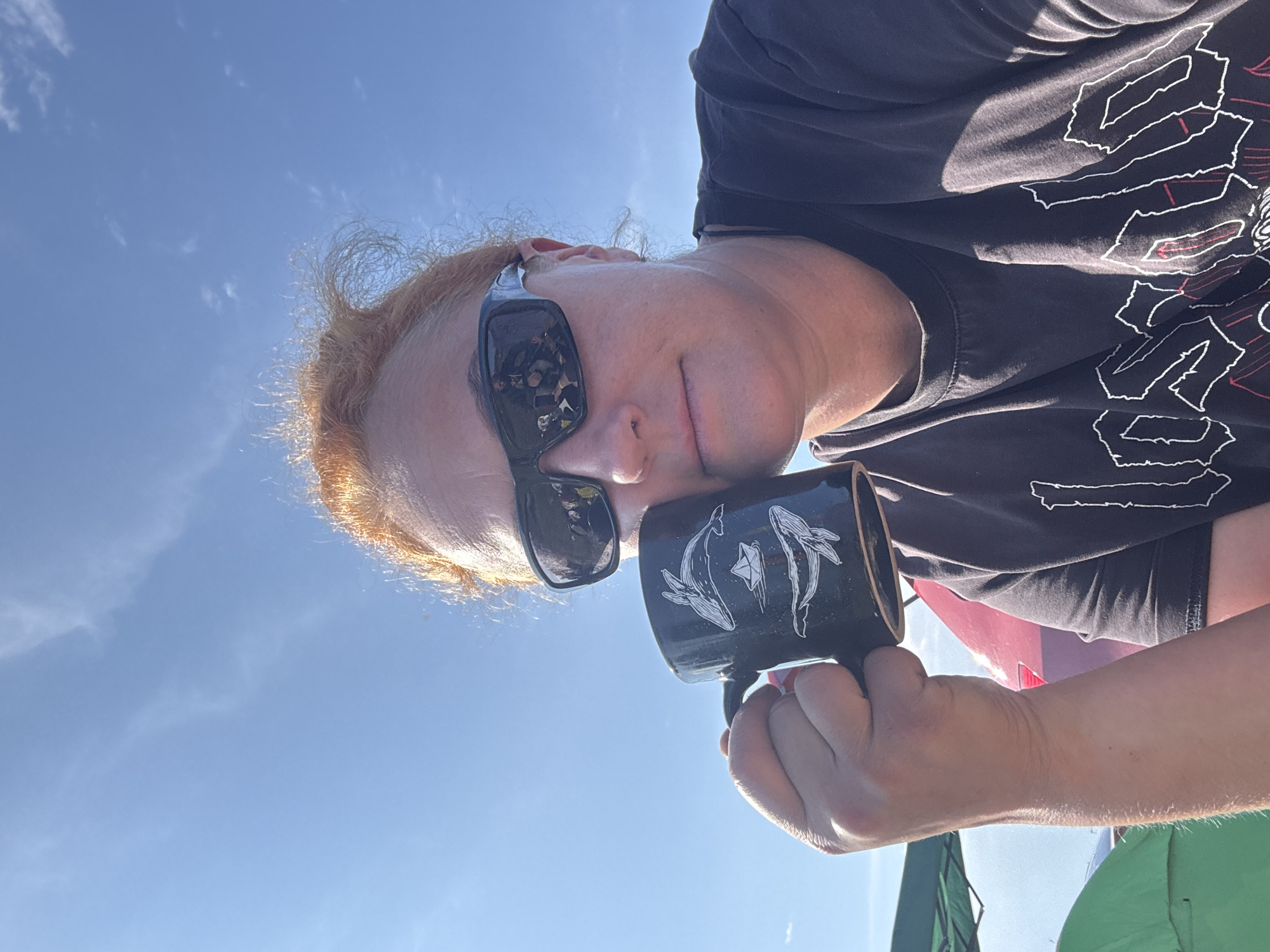 A woman with auburn hair and sunglasses is taking a selfie outdoors under a clear blue sky. She is holding a black mug decorated with white drawings of whales and a paper boat. She is wearing a dark t-shirt with a white graphic design. In the background, parts of green and red objects, possibly tents and a vehicle, are visible. The scene appears to be in an outdoor recreational area.