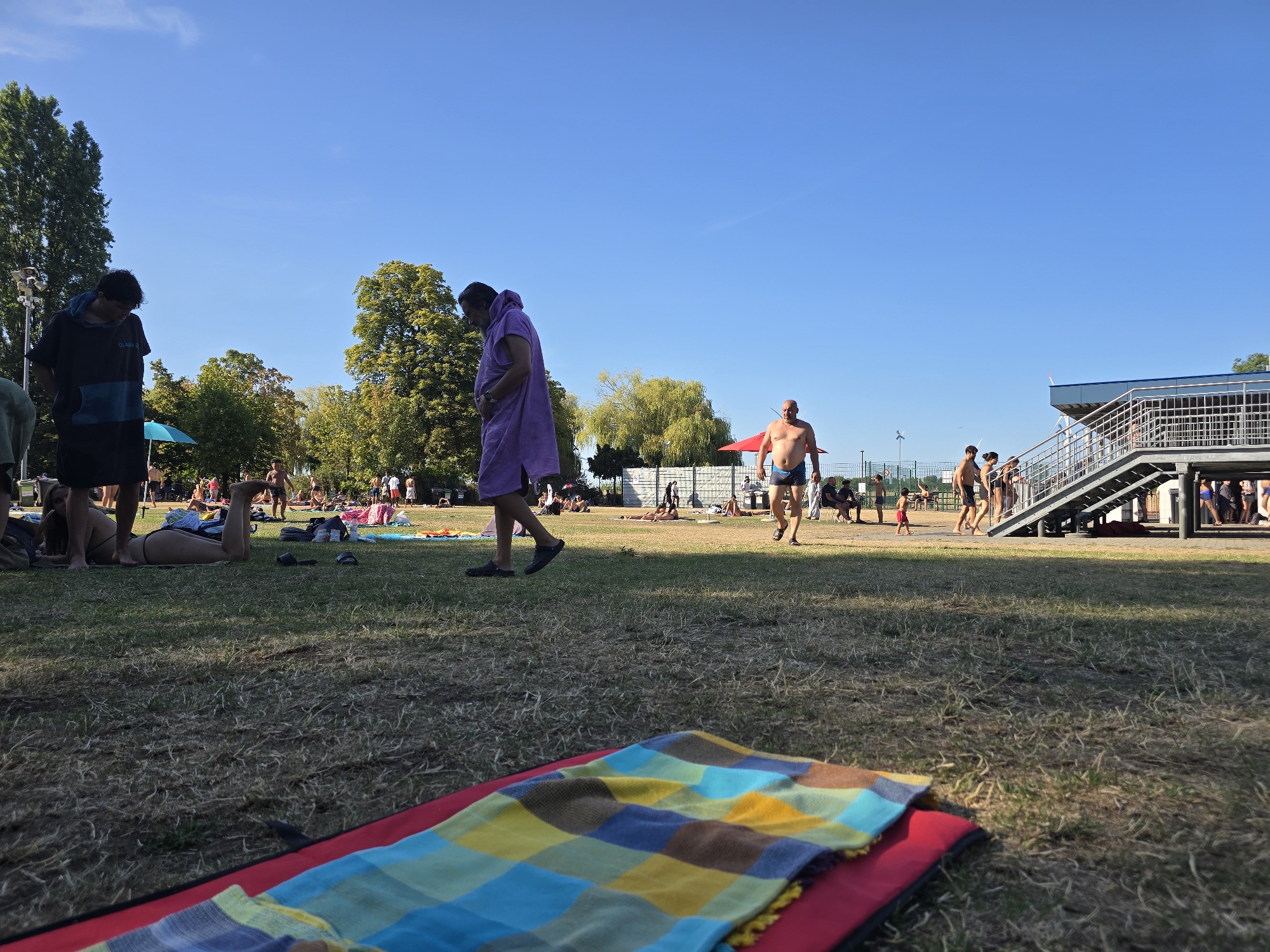 In this outdoor scene, a group of people are gathered at a public swimming pool complex on a sunny day. The foreground features a colorful checkered blanket laid out on red padding, suggesting a picnic or relaxation area. The midground shows several individuals, some dressed in swimwear and others in casual attire, relaxing on the grass or walking around. One person is wearing a purple hooded robe, while others are in bathing suits. There are trees and foliage in the background, indicating a park-like setting within or adjacent to the pool facility. To the right, a modern building with metal stairs and railings suggests the presence of changing rooms, observation decks, or entrances to the pool. The sky is clear and blue, typical of a warm summer day. This scene is consistent with the outdoor pool areas found at the Olympiadeplein in Antwerp, Belgium, a popular recreational spot known for its sports facilities and green spaces.