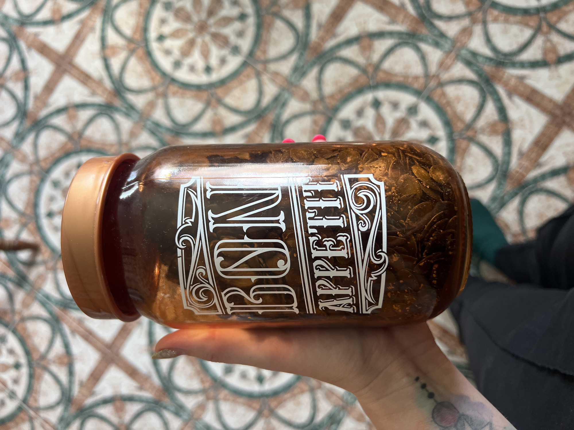 In Krasnoe-na-Volge, Russia, a hand with glittery fingernails holds a clear glass jar filled with what appears to be dried chili flakes or similar spices. The jar has a decorative white label that reads "BON APPETIT" in stylized lettering. The jar is capped with a copper-colored lid. The background is a tiled floor with a geometric pattern of brown, white, and teal.