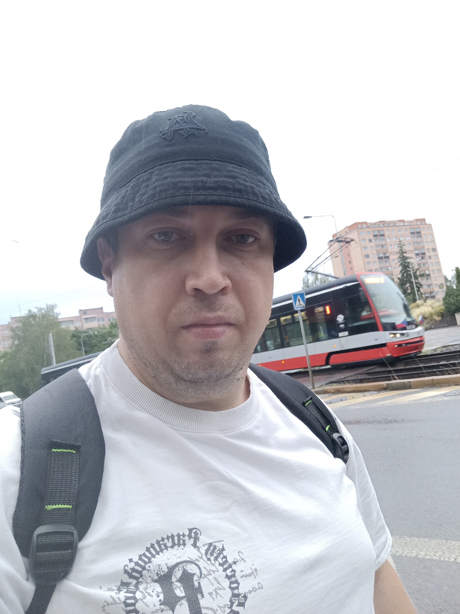 A man wearing a black bucket hat and a white t-shirt with a graphic print is captured in a close-up selfie. He has a backpack with black straps slung over his shoulders. In the background, a red and white tram is in motion on tracks, with a multi-story building and trees visible beyond. The scene appears to be an outdoor urban environment, likely a street or tram stop. The sky is overcast.