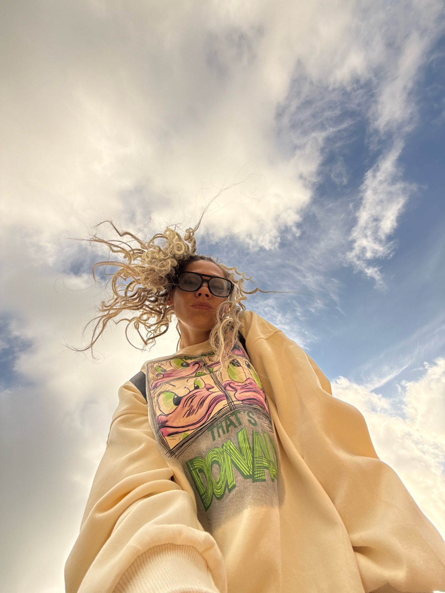 A low-angle, wide shot shows a woman from the chest up, looking up towards the camera. She has curly, blonde hair that is being blown by the wind and is wearing dark sunglasses and a light yellow sweatshirt with a graphic design on the front that includes cartoonish eyes and green lettering. The background consists of a bright blue sky filled with scattered white clouds. The image was captured outdoors, with the camera positioned beneath the subject, looking upwards. The lighting suggests it is daytime, likely sunny, with the sun illuminating the clouds from behind. The specific location in the city of Dumino, Russia, is not discernible from the image.