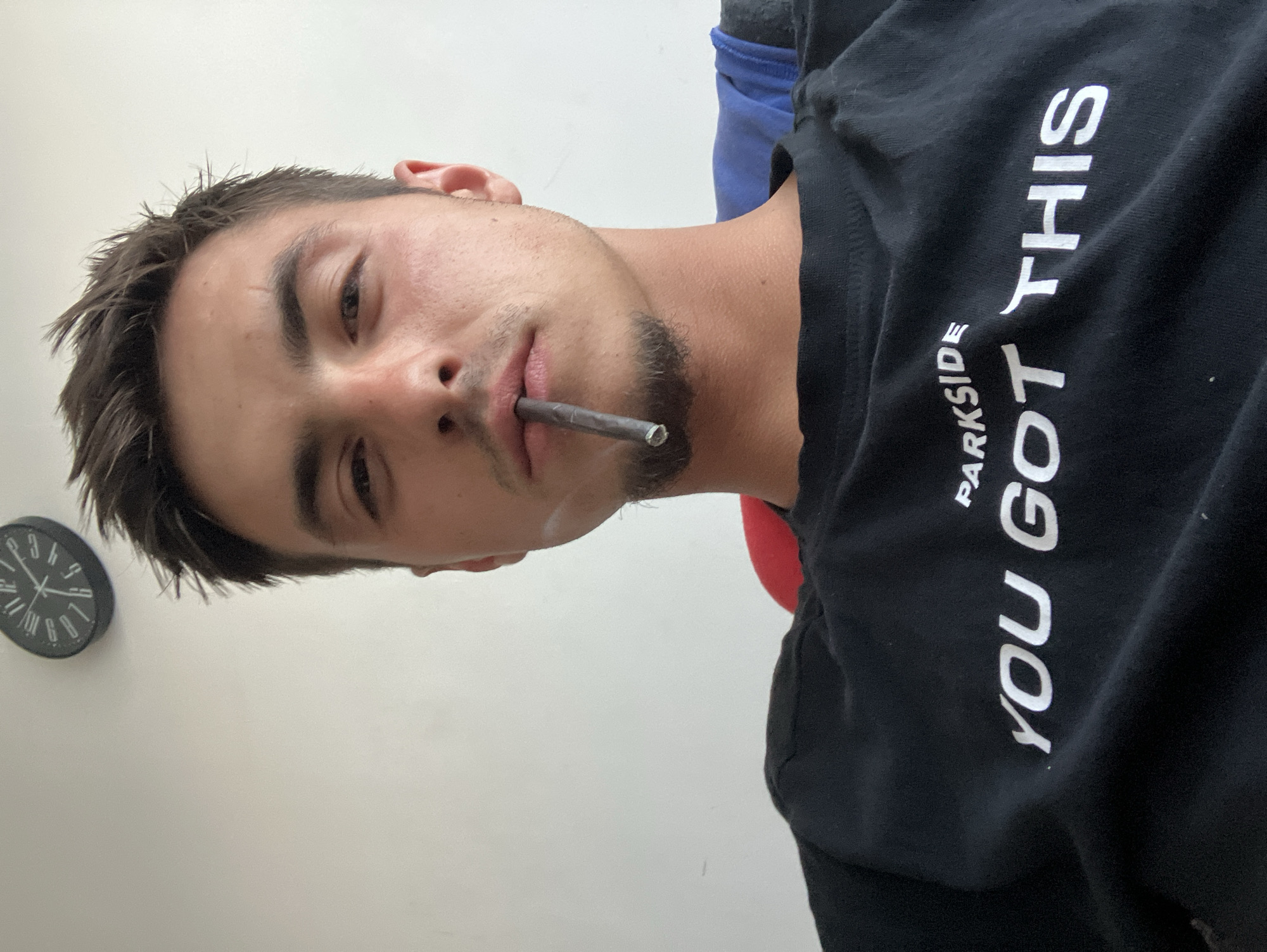 A close-up, rotated portrait captures a young man, likely in his late teens or early twenties, looking directly into the camera from a low angle. He has short, spiky dark hair, dark eyebrows, and a light mustache and goatee. A small scar or mark is visible above his left eyebrow. He holds what appears to be a lit cigarette or joint between his lips.
The man is wearing a black t-shirt that features white text. The visible words are "PARKSIDE" (smaller, above) and "YOU GOT THIS" (larger). The background consists of a plain, light-colored wall. A dark, round wall clock with white numbers is partially visible on the wall to the man's upper left. Hints of blue and red fabric are also visible behind him, possibly from clothing or furniture. The man's expression is neutral and relaxed. The scene takes place indoors. Although the location context is Plovdiv, Bulgaria, no specific visual cues within the image confirm this. The time on the clock is approximately 2:45 or 3:45, but it's hard to be precise.
