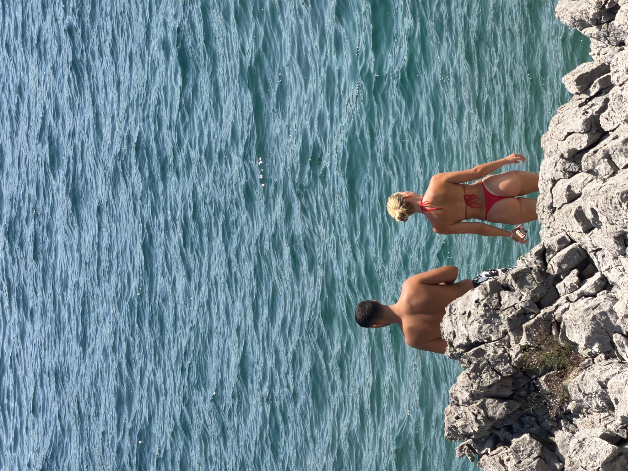 Two individuals, a man and a woman, are positioned on a rocky outcrop by the sea in Bigova, Montenegro. The man, with dark hair, faces away from the camera, looking towards the water. He is bare-chested and wearing patterned swim trunks. The woman, with blonde hair tied up, also faces away from the camera, wearing a red bikini. She holds a white object in her left hand. The sea is a deep blue-green, with visible ripples on its surface. The rocky area in the foreground is rough and textured, with some sparse vegetation growing among the stones. The lighting suggests a sunny day.