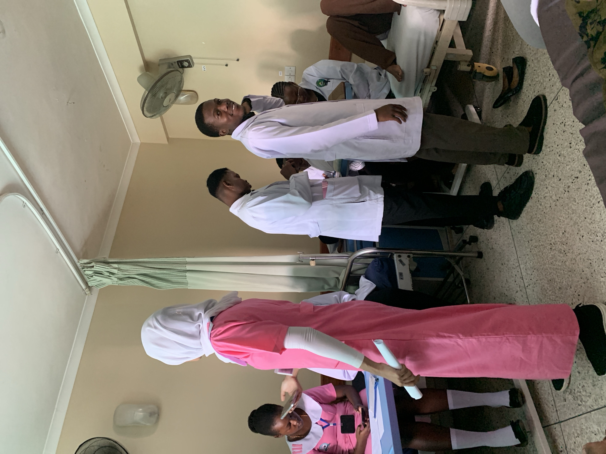 This image depicts an indoor scene within a medical facility, located in Dar es-Salaam, Tanzania. The environment is characterized by light beige walls, a white ceiling with crown molding, and a light-colored terrazzo floor. A mint-green curtain hangs from a track on the right side of the frame, alongside a wall-mounted oscillating fan and an oval-shaped wall light fixture. Another wall-mounted fan is visible on the left wall.
Eight individuals are present. In the left foreground, a female stands with her back mostly to the camera, wearing a long-sleeved pink medical scrub dress, a white hijab, and black slip-on shoes. She holds a white rolled document in her right hand. To her left, another female, dressed in a pink nurse's uniform with a white collar, white socks, and black flat shoes, is seated at a blue table, looking down at a smartphone in her hands.
In the mid-ground, two males stand, both wearing white lab coats. The male on the right faces the camera, smiling, with his left hand near his neck. He wears dark trousers and black slip-on shoes. The male to his left is partially visible, wearing a white lab coat over dark trousers and black shoes, looking downwards. Behind these individuals, a metal hospital bed frame is partially visible.
In the background, other individuals in white lab coats are present. One is partially obscured by the standing males, while another is seated near a hospital bed. To the far right, part of a hospital bed with white linens is visible, occupied by a patient whose bare lower legs and feet are resting on the bed. A small, dark yellow medical device or shoe is on the floor near the patient's feet. An IV pole is partially visible between the patient and the standing males.