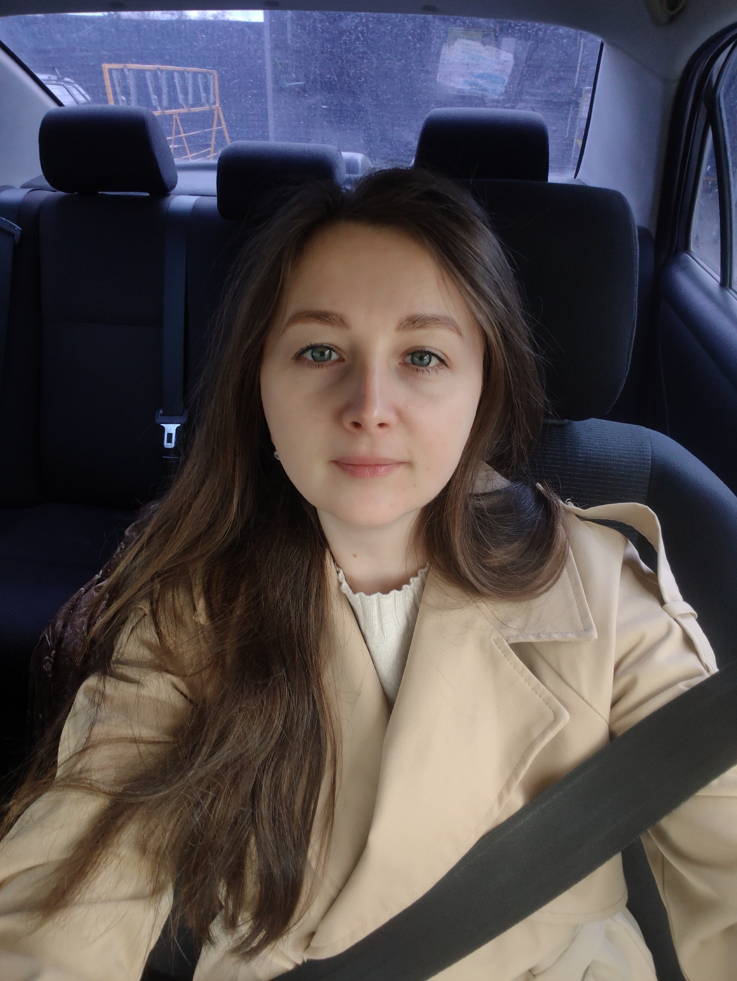 A woman with light skin and long brown hair, wearing a beige trench coat and white top, is seated in the back of a car. She is wearing a seatbelt. The interior of the car is dark, with black seats and headrests. Outside the rear window, a yellow barrier is visible through a hazy or rain-streaked window. The location appears to be urban, but specific details are unclear due to the window's condition.