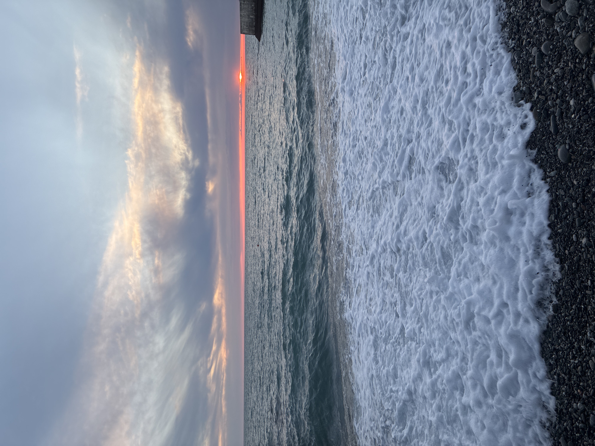 This is a photograph of a pebble beach and the sea at sunset in Sochi, Russia. The sky is filled with wispy clouds tinged with orange and pink from the setting sun, which is partially visible on the horizon to the right. The sea is a deep teal color with gentle waves breaking on the shore, creating white foam. In the distance, a concrete structure, possibly a pier or breakwater, extends into the water. The beach itself is covered in smooth, dark pebbles. There are no people visible in the image.