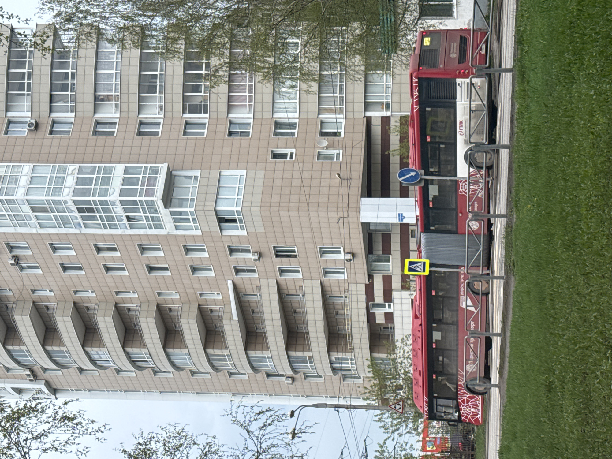 A high-rise apartment building with multiple balconies stands next to a red bus in Perm, Russia. The bus is partially obscured by a metal fence, next to a grassy area. Traffic signs indicating pedestrian crossing and directions are visible. Trees are scattered near the building and along the sidewalk.