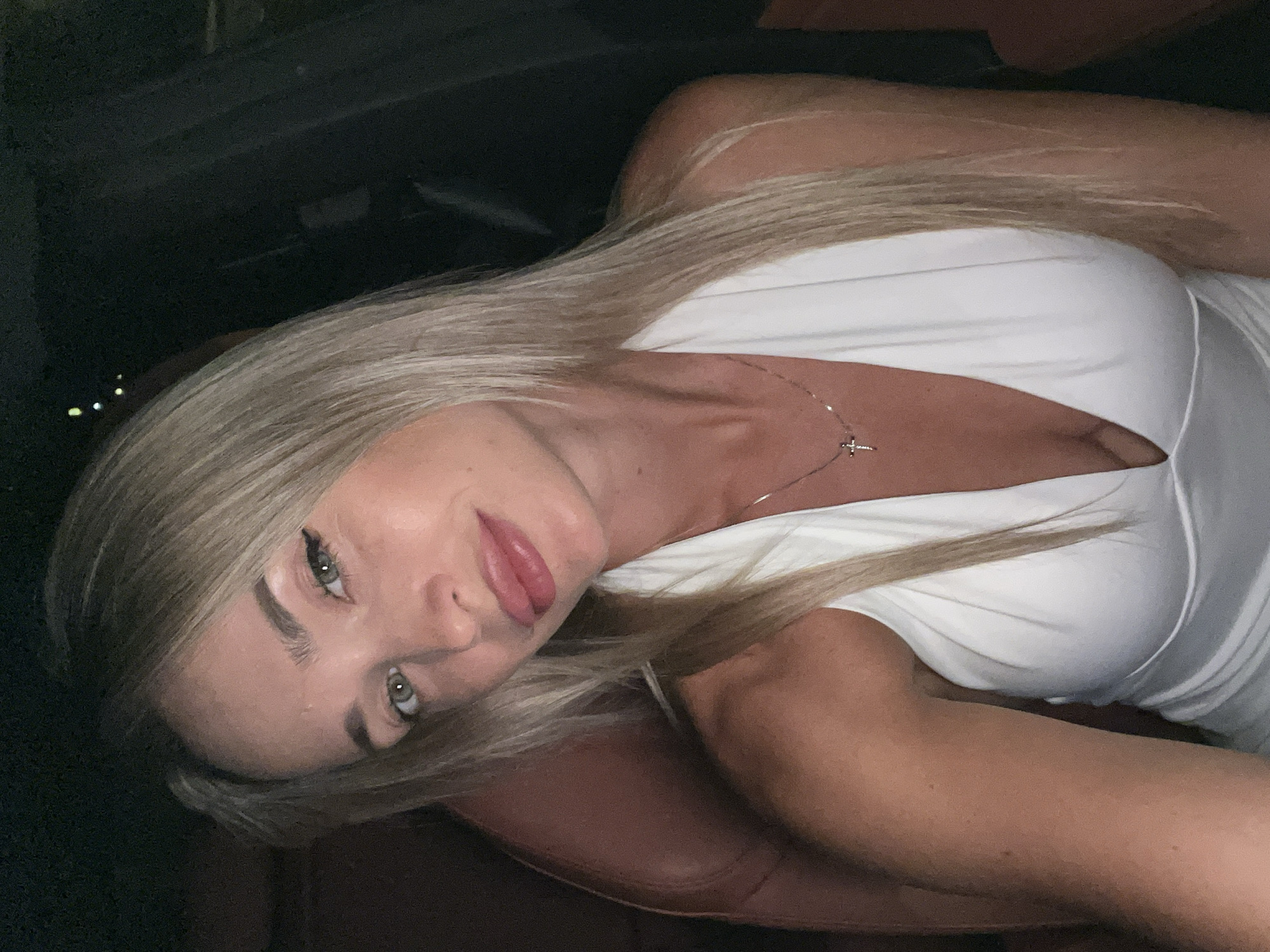 A woman with blonde hair and green eyes is pictured in a car, at night. She is wearing a white dress with a deep V-neckline and a silver necklace with a cross pendant. Her skin appears to be tanned. The background is dark, suggesting it is nighttime. The interior of the car appears to be leather-upholstered. The image was taken in Sharm El Sheikh, Egypt.
