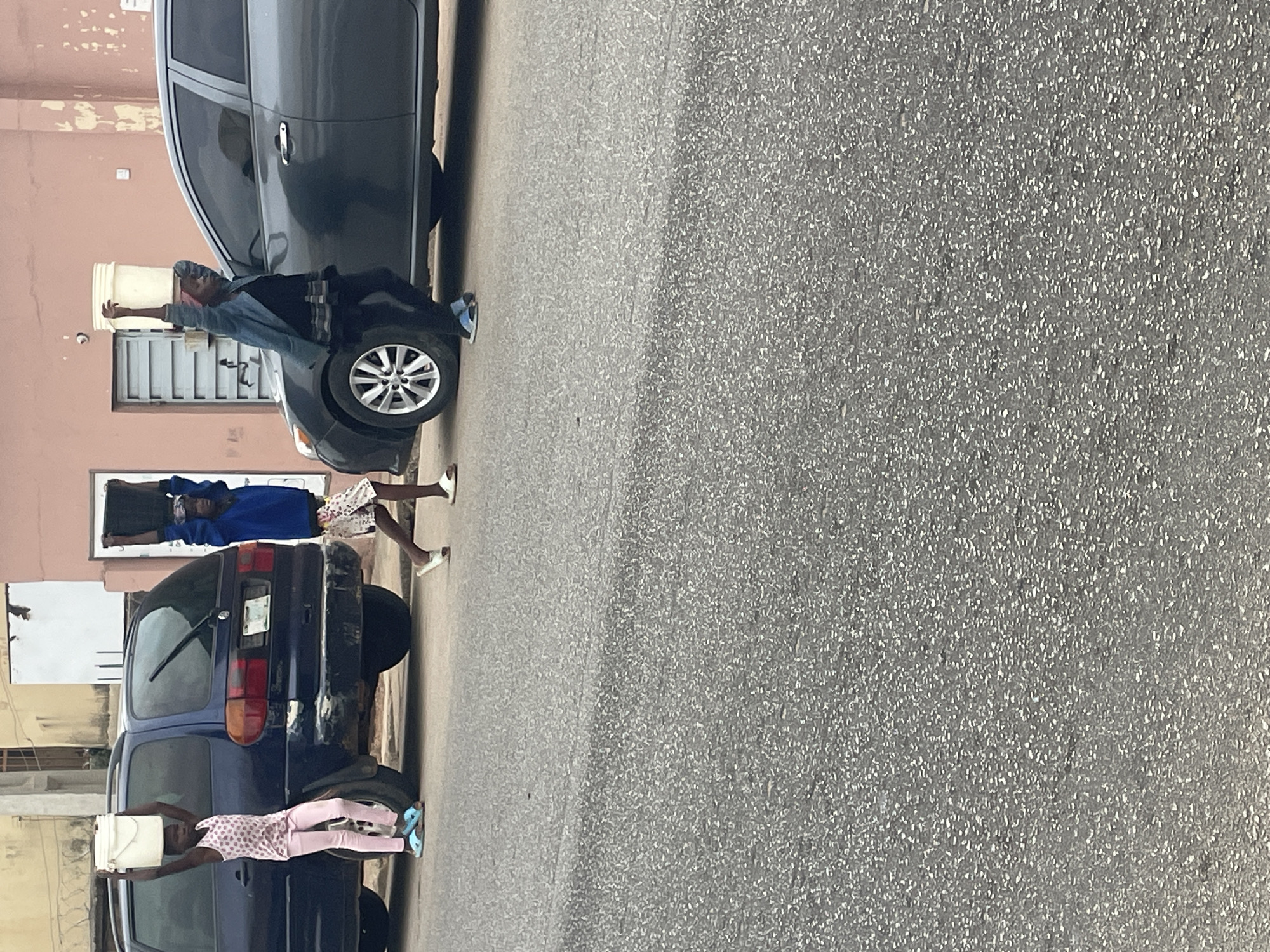 The image shows three children carrying buckets on their heads in Jos, Nigeria. The children, dressed in casual clothing, are walking along a street adjacent to parked cars. The street is paved. The buildings in the background appear to be residential. The image is taken during daylight.