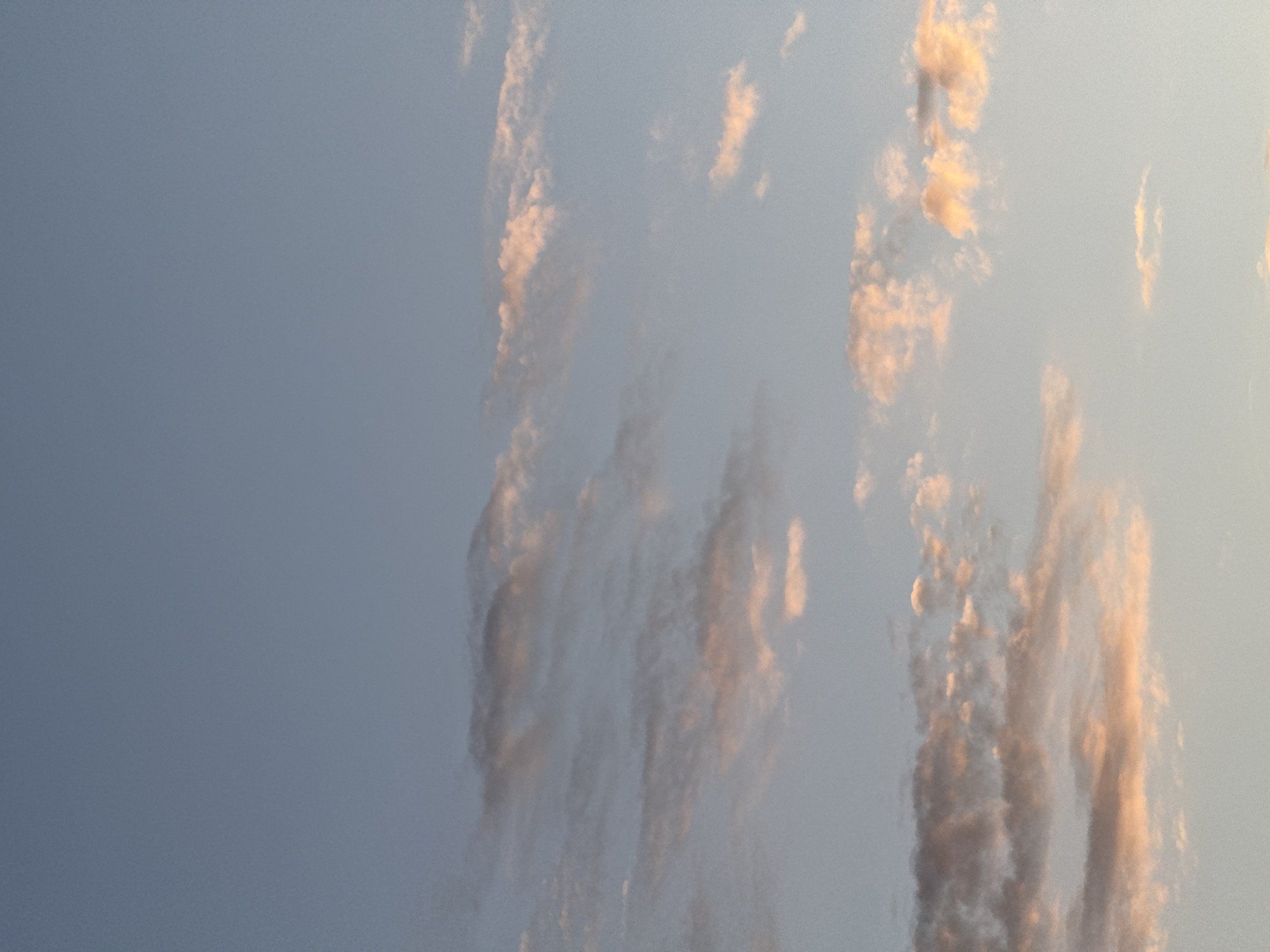 The image shows a section of the sky, possibly in Мокино, Russia. The left side is a plain, light blue gradient, indicating clear sky. On the right, there are streaks of elongated, textured clouds with a gradient of light pink/orange hues, potentially from the sun. The clouds appear vertical.