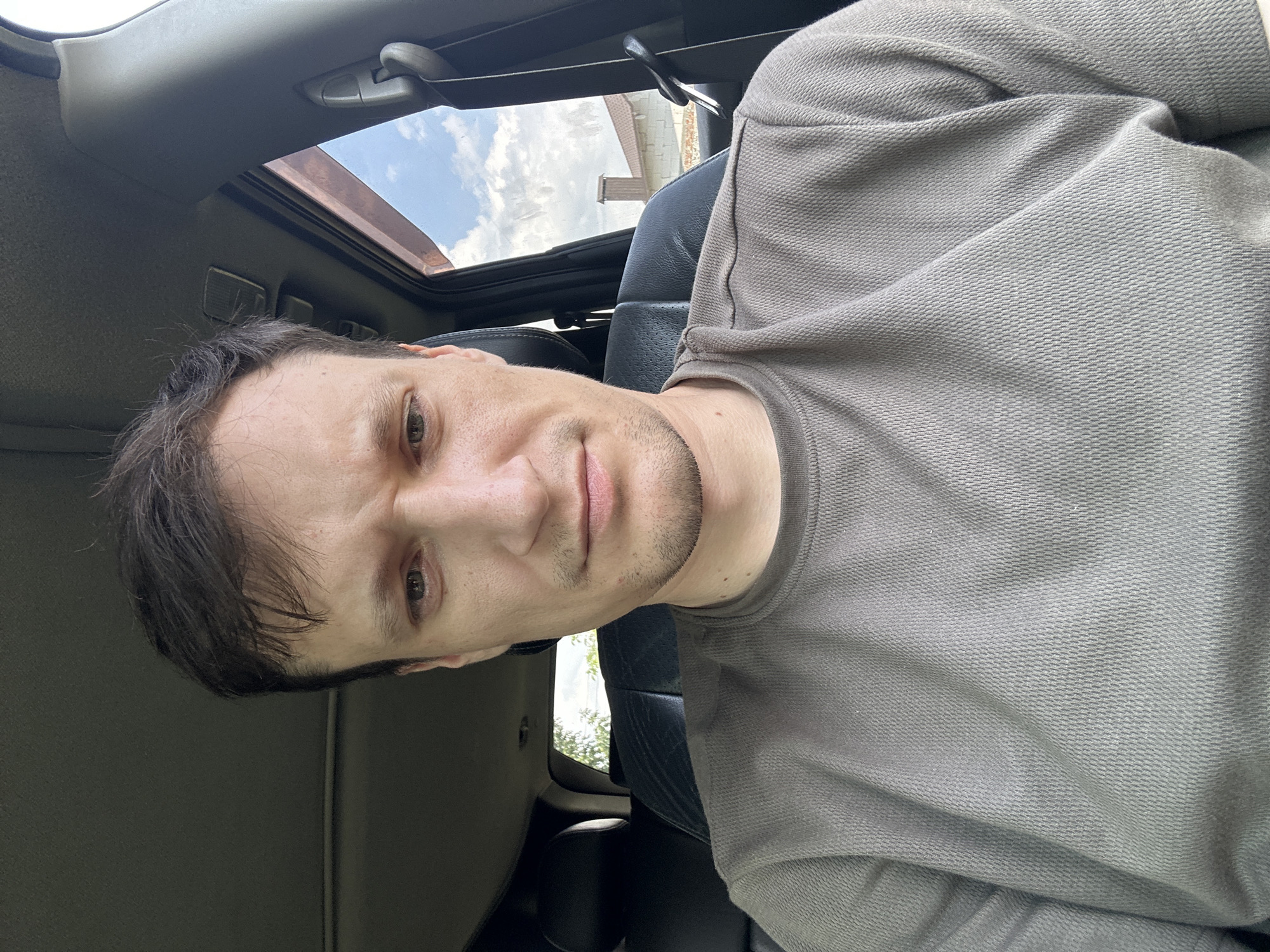 A man is sitting in the driver's seat of a car, looking directly at the camera. He has short, dark brown hair, fair skin, and a light stubble on his chin and upper lip. His eyes are brown, and he has a furrowed brow, suggesting he might be concerned or concentrating. He is wearing a plain, taupe-colored t-shirt. The interior of the car is visible, including the black leather driver's seat and a portion of the car's headliner. Outside the car window, the sky is cloudy and blue, with a glimpse of a house with a brick chimney and part of a fence. The location is identified as Makiv, Ukraine.