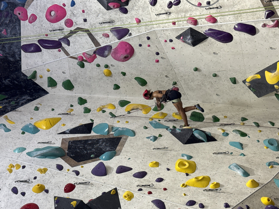 An indoor climbing gym in Bangkok, Thailand, features extensive, multi-faceted walls constructed from light grey panels and geometric black sections. The walls are densely equipped with an assortment of climbing holds in diverse shapes and sizes, colored yellow, teal, green, purple, pink, and red. A single individual, appearing female, is observed in a controlled descent on a vertical wall section. She is equipped with a dark sports bra, dark shorts with a light trim, a red bandana on her head, and climbing shoes. Secured by a climbing harness, she is attached to a prominent green climbing rope. Her hands are positioned near her waist, actively managing the rope for her descent. Her left foot is firmly placed on a large, black, multi-faceted climbing volume, while her right foot maintains contact with the light grey wall. Multiple quickdraws are mounted on the wall's anchor points, and additional climbing ropes hang vertically throughout the structure.