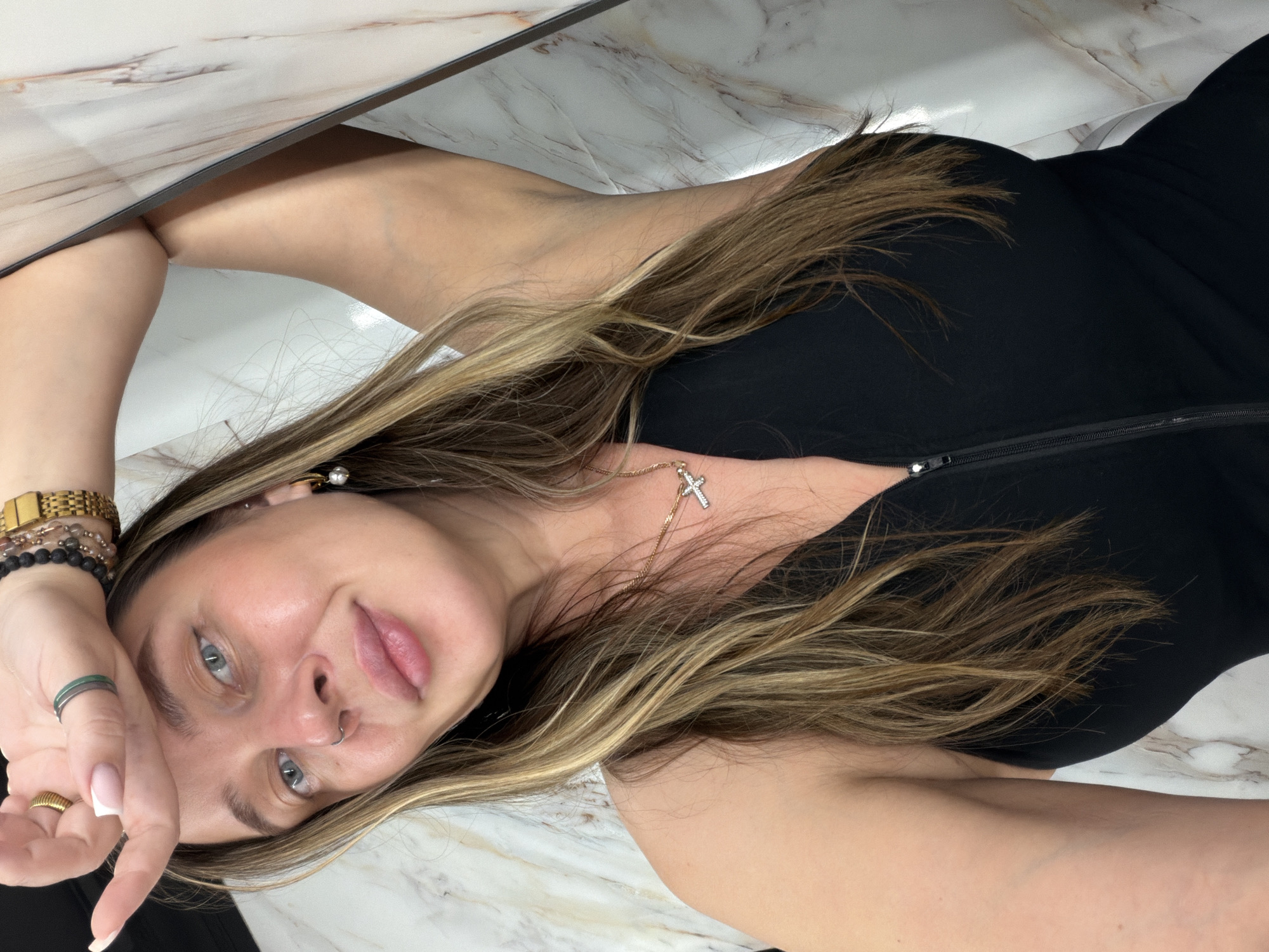A woman with blonde highlighted hair is lying on a white and beige marble-patterned surface. She is wearing a black top and a gold necklace with a cross pendant. Her left hand is raised to her forehead, adorned with multiple rings, bracelets, and a gold watch. Her face is visible, showing light blue eyes and a nose ring. Based on the architectural details visible in the image, there is no indication that the photo was taken in Moscow, Russia. The location cannot be determined from the available information.