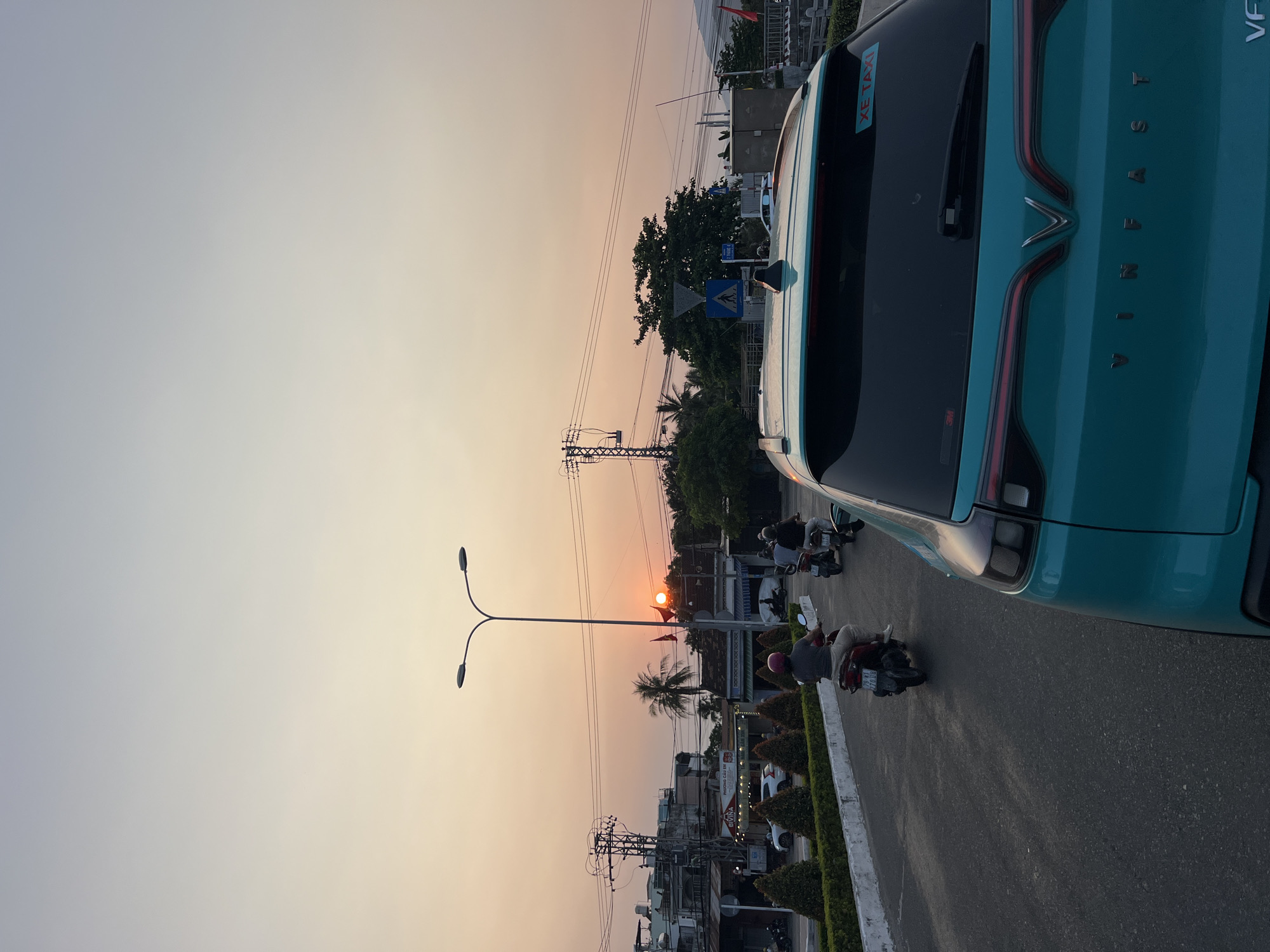 The image captures a sunset scene in Nha Trang, Vietnam. The sky displays a gradient of light orange near the horizon, fading into a pale blue-gray further up. A VinFast car is prominently featured on the right, its teal color distinct. The word "VINFAST" is visible on the car's rear. On the road to the left, several individuals are riding motorbikes. The street is lined with green shrubbery and a concrete barrier. In the background, there are buildings, utility poles with power lines, and palm trees silhouetted against the setting sun. The sun appears to be directly above the overhead street lighting pole. A "XE TAXI" sticker is present on the VinFast vehicle. The area seems like a typical urban environment with a mix of traffic and infrastructure, common for Vietnamese cities.