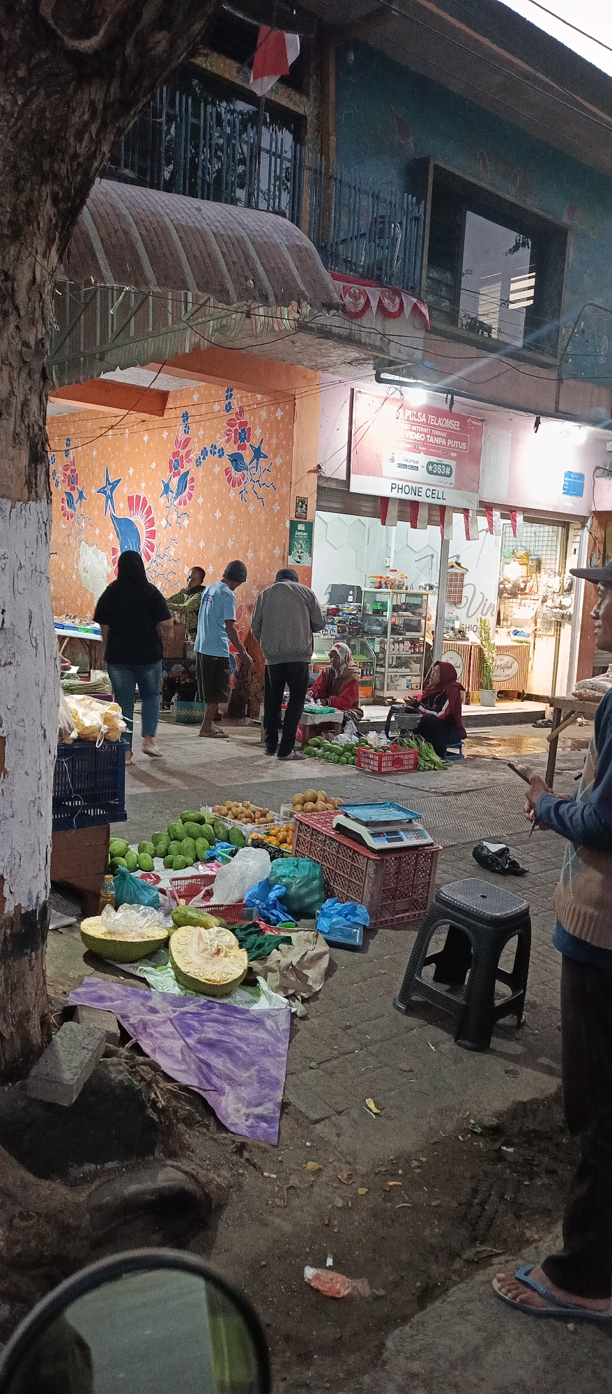The image depicts an outdoor market scene in Ardirejo, Indonesia, likely during the late afternoon or early evening, given the lighting conditions. In the foreground, a variety of fruits and vegetables are displayed on the ground, including what appears to be green mangoes, yellow fruits, and a large, cut jackfruit. Various crates, bags, and a weighing scale are scattered amongst the produce.
Several individuals are present. To the left, a person in a black hijab and dark pants stands near a table with more produce. Two men, one in a blue t-shirt and shorts, and another in a grey jacket and dark pants, are engaged in conversation near a stall. Further to the right, two women are seated, possibly vendors, attending to their goods. One woman is wearing a red headscarf and red jacket, and the other is wearing a maroon hijab and jacket. A black plastic stool is positioned near them.
In the background, there is a building with an orange-painted wall adorned with floral patterns. A shop front with a sign advertising "PHONE CELL" is visible, suggesting a commercial area. Red and white Indonesian flags are displayed on the building, indicating a national celebration or event. The ground is paved with an uneven surface, with some dirt and debris present. A motorcycle's side mirror is partially visible in the bottom left corner, suggesting the photographer is on a motorcycle. The overall atmosphere appears to be that of a bustling local market.