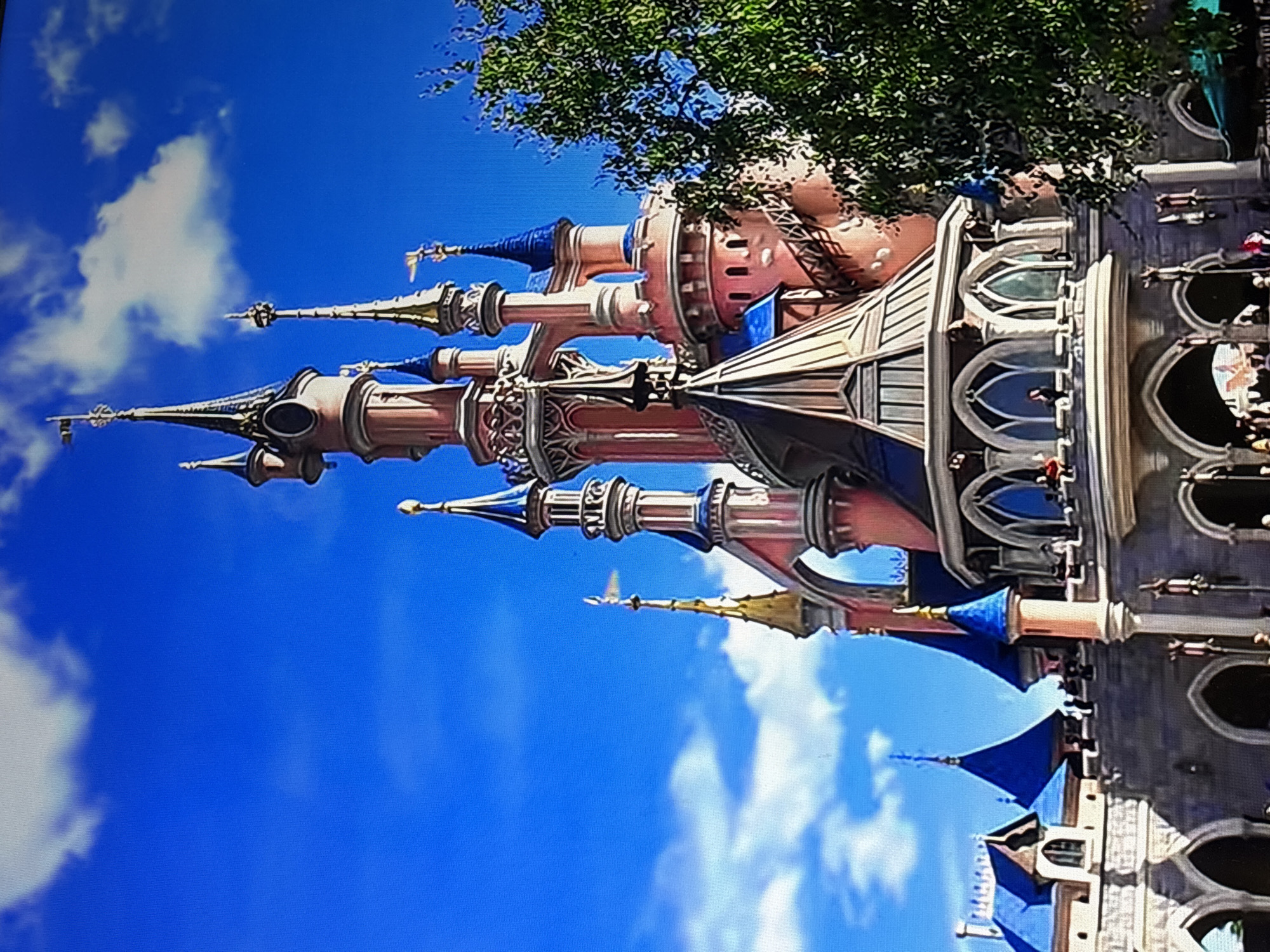 The image shows Sleeping Beauty's Castle at Disneyland Paris in Marne-la-Vallée, France. The castle's pink and blue spires are visible against a blue sky with scattered white clouds. A tree partially obscures the top of the castle. People can be seen on walkways or balconies within the castle structure. The photograph is oriented sideways.