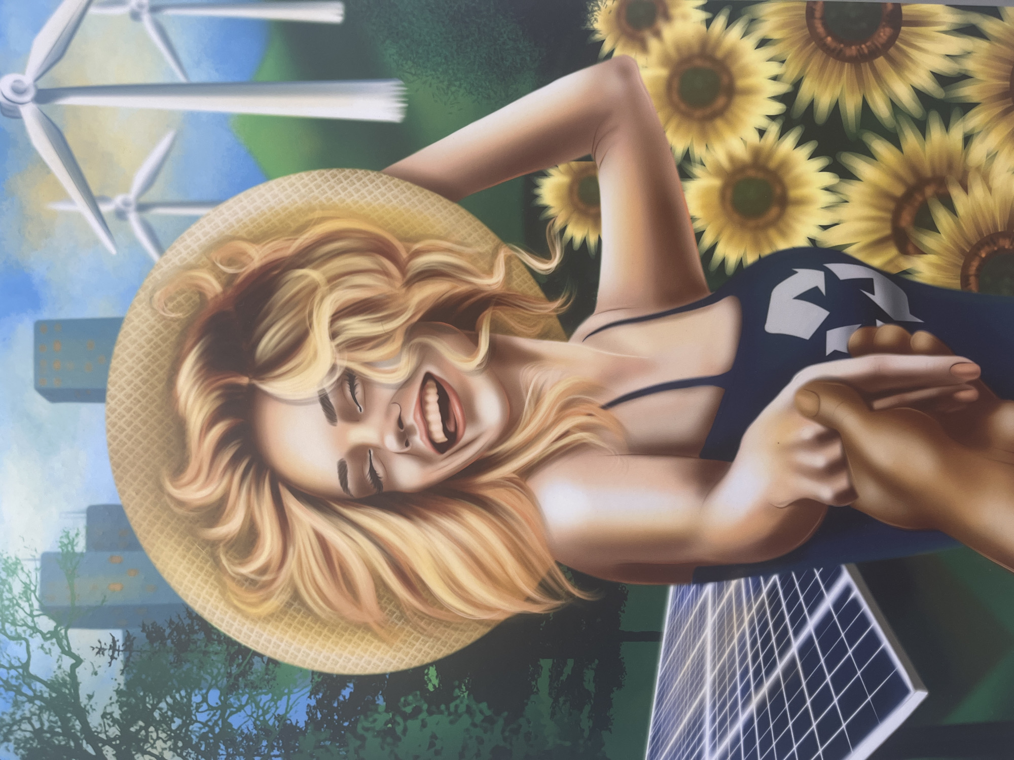 A woman in a sun hat lies on her back wearing a dark-colored tank top with a recycling symbol on it, and is holding someone's hand. She is laughing with her eyes closed. Behind her are several sunflowers and a solar panel. In the background are wind turbines and the suggestion of a city landscape with green spaces, possibly representing the city of Varna, Bulgaria.