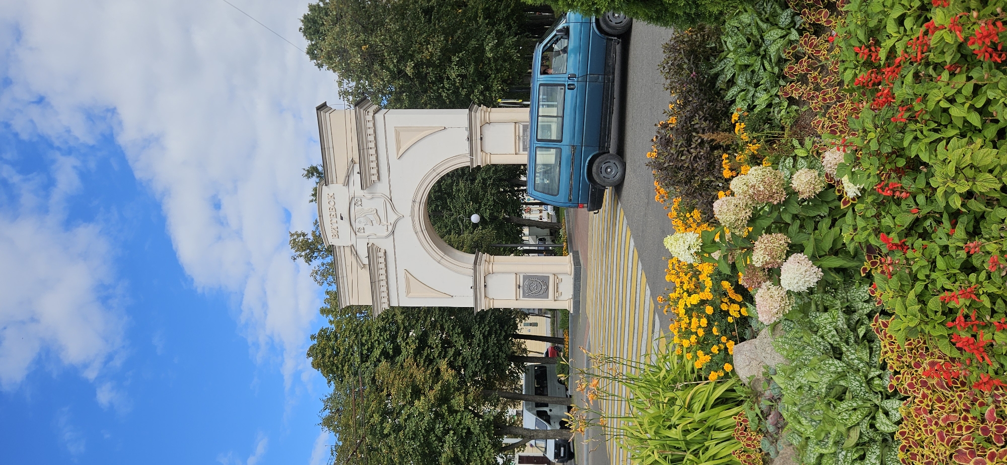 The image shows a view of the Victory Arch in Vitebsk, Belarus, on a partly cloudy day. The arch, a prominent city landmark, is made of light-colored stone and features intricate detailing and inscriptions. A blue minivan is visible driving under the arch, and a white van is parked to the left. In the foreground, a vibrant flower bed bursts with a variety of colorful blooms, including yellow marigolds, pink and white hydrangeas, and red salvia, along with lush green foliage. A pedestrian crossing with yellow and white stripes is also present. The background consists of mature trees and a bright blue sky with scattered white clouds.