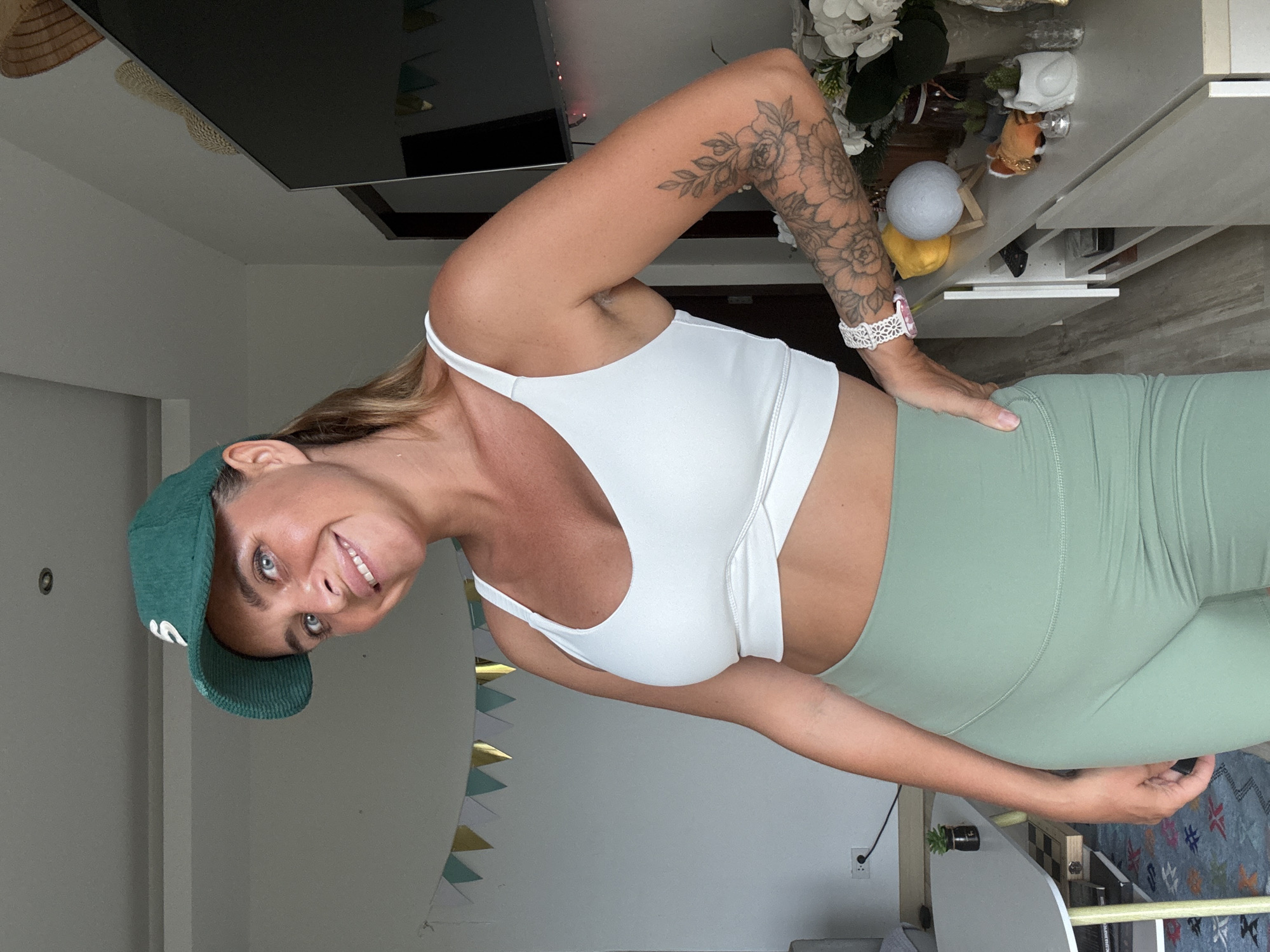 A woman with blonde hair pulled back and a green baseball cap is standing in a room. She is wearing a white sports bra and green shorts. She has tattoos on her left arm. The room appears to be a living area, with a TV on a stand in the background. There are decorative items on the stand, including a lamp and some small figurines. A table with a chessboard is visible to the left. The overall lighting suggests it is daytime. The scene is likely indoors, possibly in a residential setting within the city of Ko Samui District, Thailand, given the provided context.