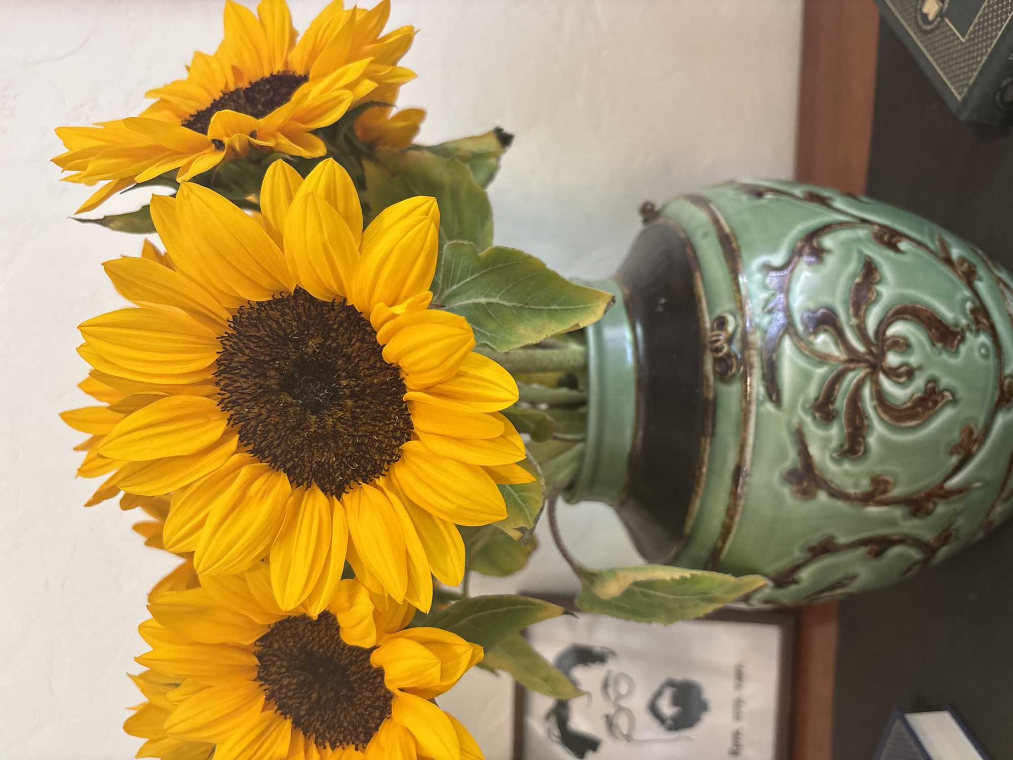 The image shows a vase of sunflowers. The vase is light green with a brown floral design. It sits on a dark surface, possibly a shelf or table, with a framed image visible behind it. The sunflowers are bright yellow with dark brown centers. The image appears to be taken indoors, with a white wall visible in the background. The location in the city of Nha Trang, Vietnam is not evident from the image.