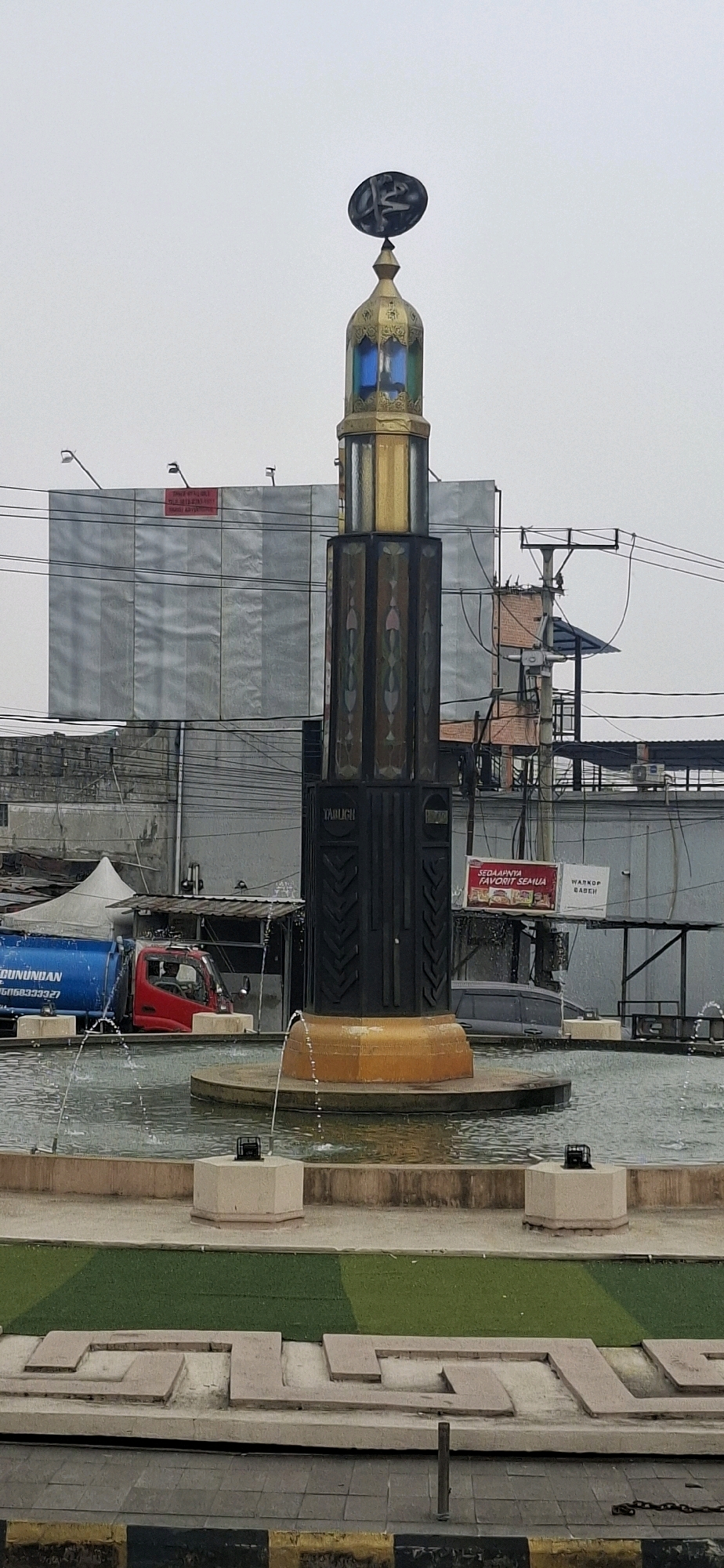 The image depicts the Tajug Rengganis Monument in Cianjur, Indonesia. The monument, a tall, dark structure with decorative panels and a golden top, stands in the center of a fountain. The monument features an Islamic calligraphy symbol at its apex, set against an overcast sky. Around the fountain, there are fountains spraying water. In the background, a large building covered with a silver tarp and a red sign is visible, along with power lines, a red truck with a blue water tanker, and other structures, suggesting an urban environment. The foreground shows a patterned pathway and landscaping.
