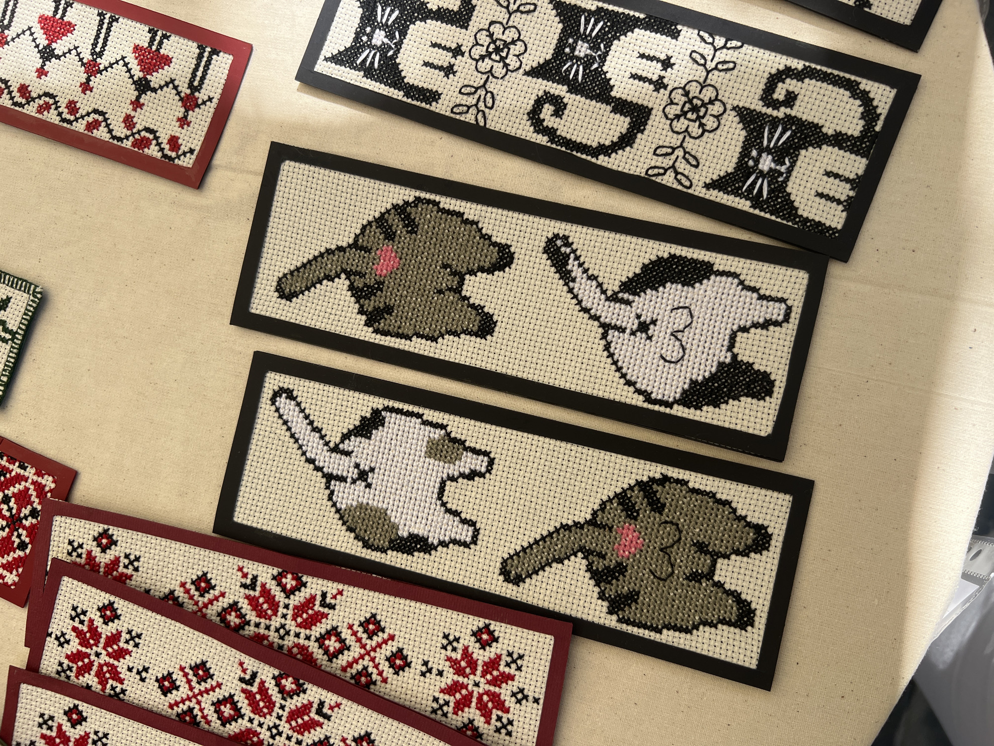 The image displays a collection of cross-stitched items, primarily bookmarks, laid out on a beige surface, likely a table or fabric. The items feature various designs, including traditional red and black floral patterns on some, and humorous cat-themed designs on others. Specifically, there are several bookmarks depicting the rear ends of cats, some with a pink heart. Other bookmarks show full-body black and white cats with floral accents. The lighting suggests an indoor setting, possibly a craft fair or a home studio. The scene is static, with no people or actions depicted. The location in Chișinău, Moldova cannot be determined from the visual information provided.