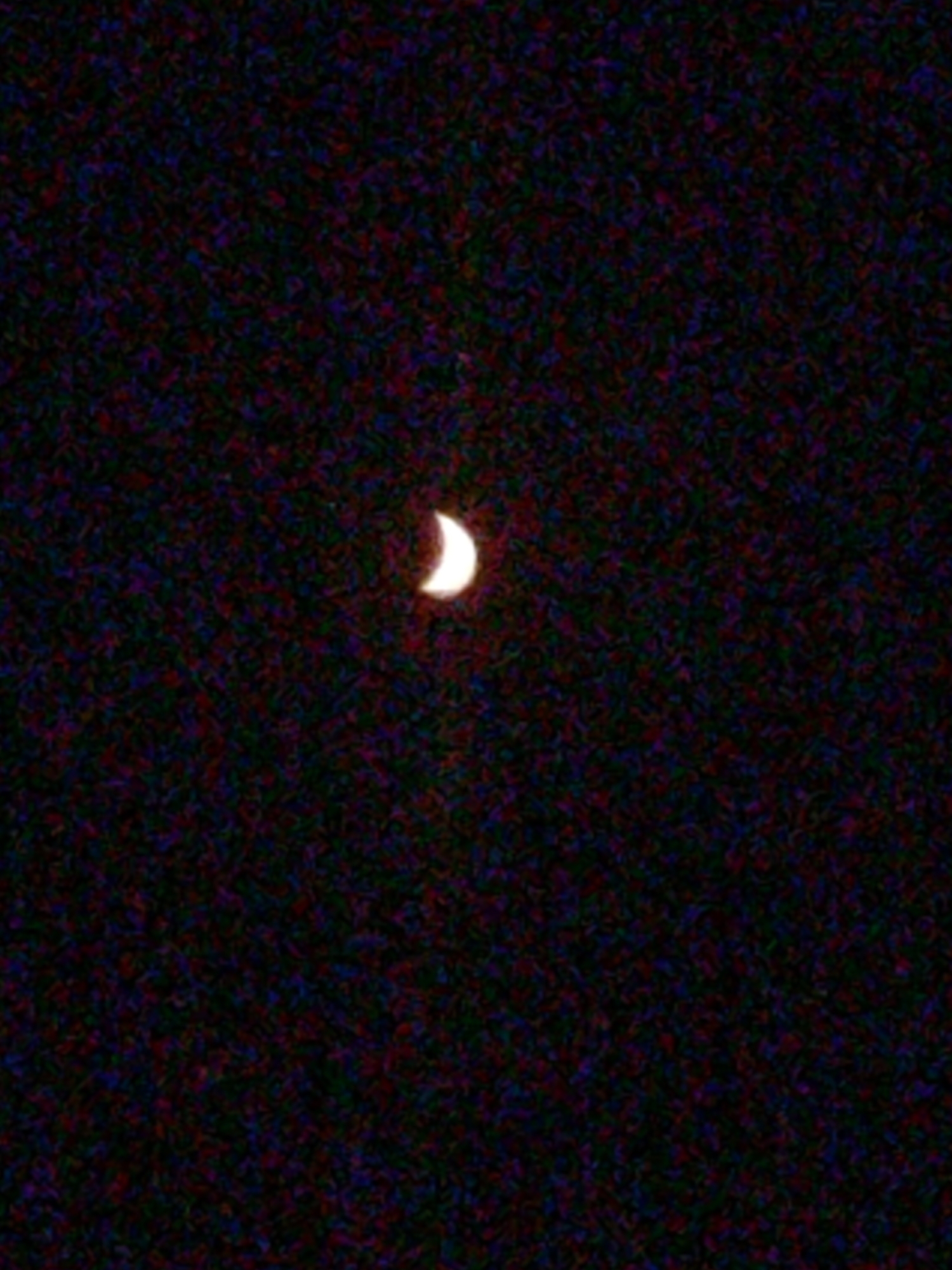 The image shows a crescent moon in a dark, grainy sky. The moon is positioned in the upper center of the frame and is illuminated on its left side. The background is filled with noise, appearing as a textured mix of dark colors, primarily black, with hints of red, blue, and green. There are no other discernible objects or features in the image.