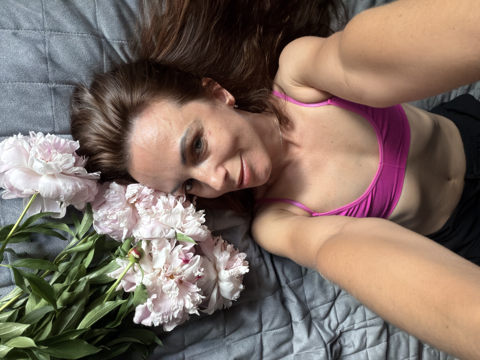 A woman with long, dark brown hair lies on a gray, quilted surface, possibly a bed or couch. She is wearing a bright pink sports bra. In the lower left quadrant of the image are several light pink peonies with green leaves. The woman is looking towards the camera with a slight smile. Her right arm is extended, and it appears she is taking the photo herself, which is a selfie. The lighting suggests it is daytime, possibly indoors due to the lack of strong shadows. The overall mood is relaxed and serene. There is no visible text in the image. The location context of Birobidzhan, Russia, does not provide any direct visual cues within the image itself.