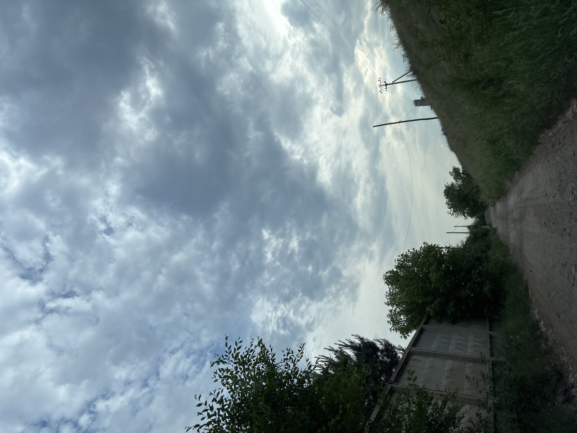 The image depicts a dirt road leading into the distance, bordered by a concrete fence on the left and a grassy, sloped embankment on the right. The sky is heavily overcast with a mix of light and dark gray clouds. Several trees are visible along the road, with denser foliage on the left side near the fence and sparser vegetation on the right. Electrical poles and wires are present, with one pole more prominently featured on the right side, and a chimney-like structure is visible in the distance on the embankment. The overall scene suggests an outdoor, possibly rural or industrial, setting within the city of Ust-Abakan, Russia. There are no people visible in the image.