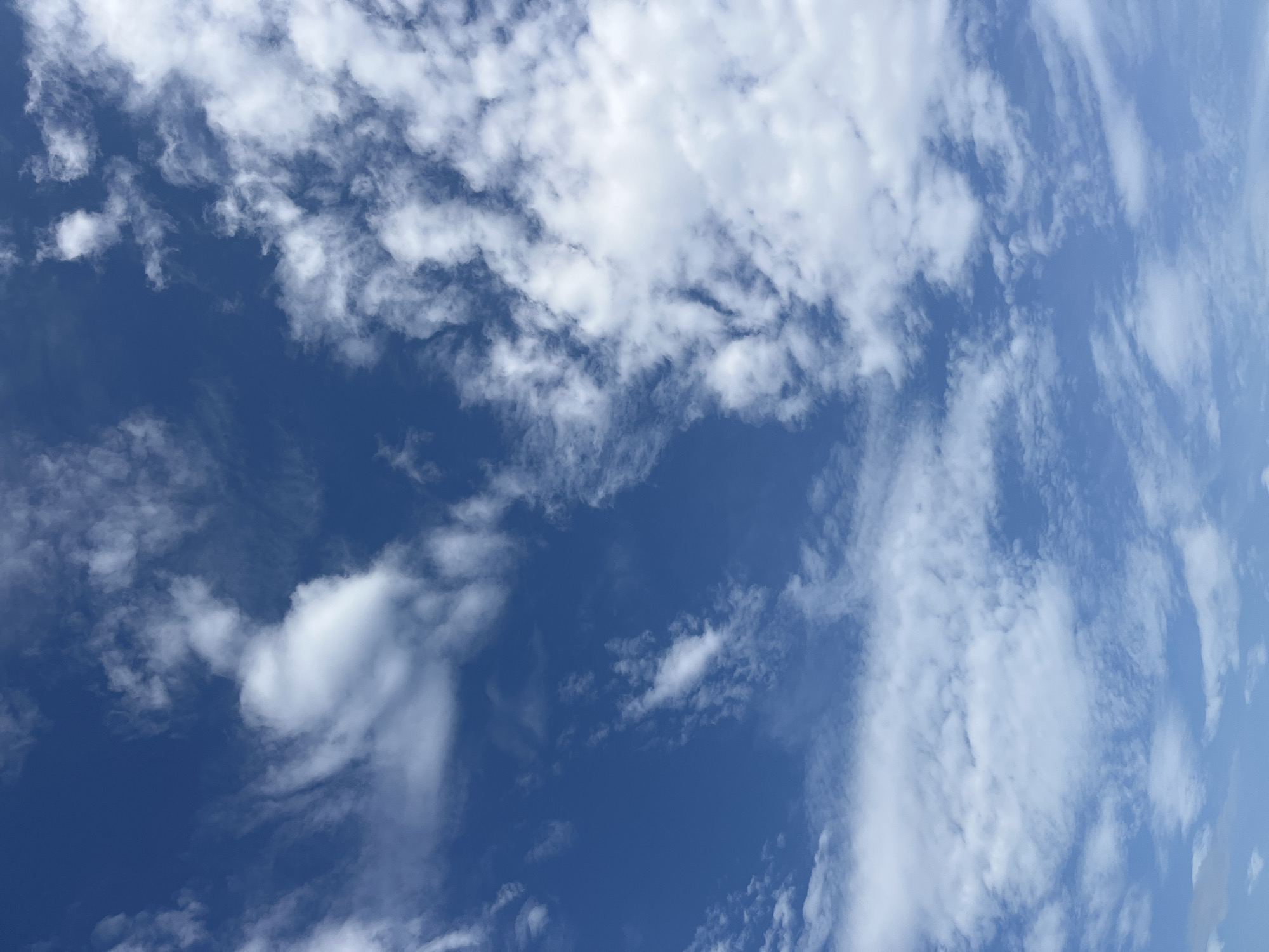 The image captures a bright blue sky filled with various white clouds. The sky exhibits a rich, deep blue hue, particularly in areas less covered by clouds. The clouds themselves are diverse in shape and density, ranging from soft, wispy streaks and patches to more substantial, fluffy formations.
In some sections, the clouds appear thin and elongated, suggesting cirrus or high-altitude stratocumulus types, while other areas feature denser, more scattered cumulus clouds with defined edges, some casting subtle shadows on themselves. The cloud cover is uneven, with significant portions of clear blue sky visible between the white formations. The lighting suggests it is daytime, likely a bright and sunny day, with the sun illuminating the clouds from above, making them appear brilliant white against the blue backdrop. There are no visible objects, people, or text in the image. The scene is purely a depiction of the sky and its cloud patterns.