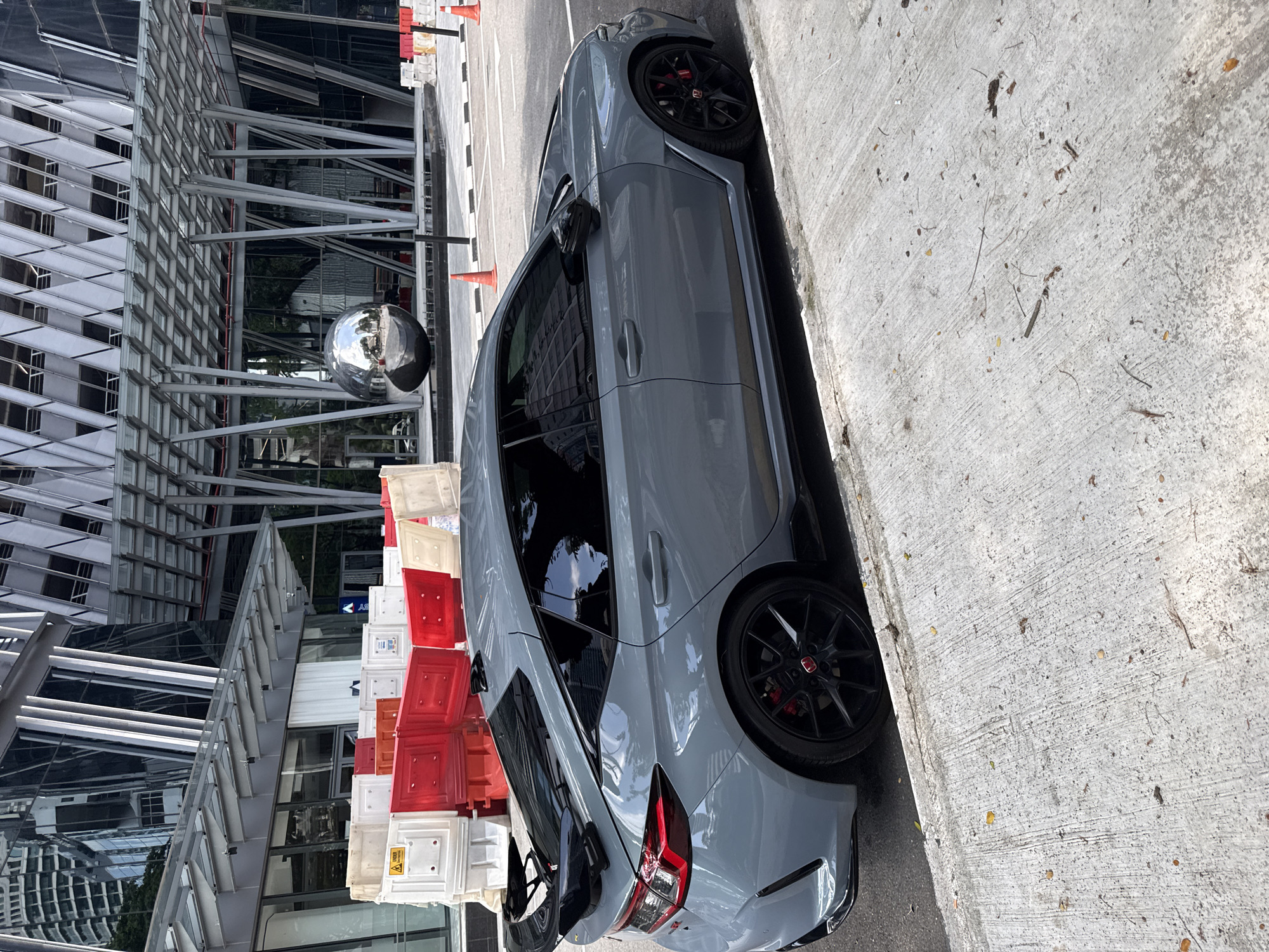 The image shows a parked grey Honda Civic Type R in an urban environment. The car is positioned on the right side of the frame next to a concrete curb. Behind the vehicle, there are red and white traffic barriers and a modern building with a reflective orb-like sculpture in front. The architecture suggests a contemporary design with metal frameworks and glass panels. The location appears to be in Kuala Lumpur, Malaysia, evidenced by the architectural style and urban infrastructure. There are no visible people in the frame.