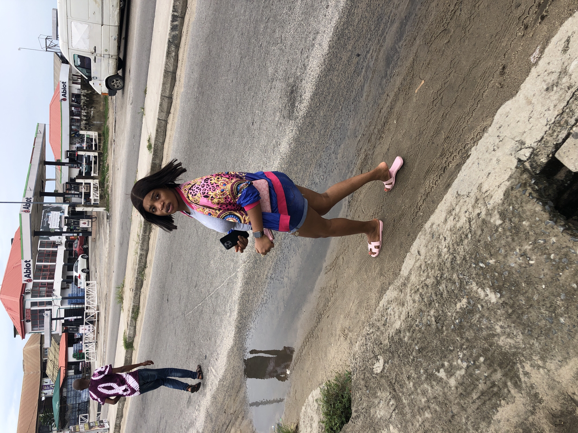 The image shows a young woman standing near a large puddle in Ado, Nigeria, holding a cell phone, likely capturing a reflection of herself. She is wearing a colorful shirt, denim shorts, and pink sandals. She is standing on a dirt shoulder of a concrete sidewalk next to a paved road. In the background are several buildings. To the left is a man walking in the opposite direction. An Abiot gas station is visible across the road. A white van can be seen parked on the road.
