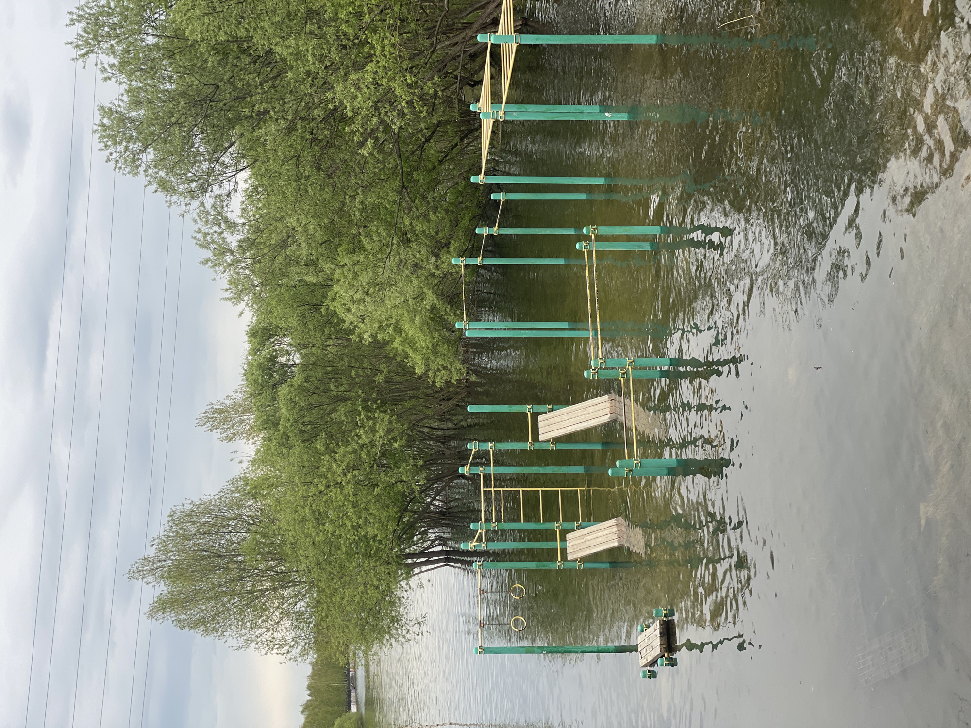 The image depicts a flooded outdoor fitness area in a park. The fitness equipment is green and yellow and partially submerged in murky water. The water level appears to be high, obscuring the ground beneath. Trees with green leaves line the edge of the water. Overhead power lines are visible in the background. The location is Krasnoyarsk, Russia. There are no people visible in the image.