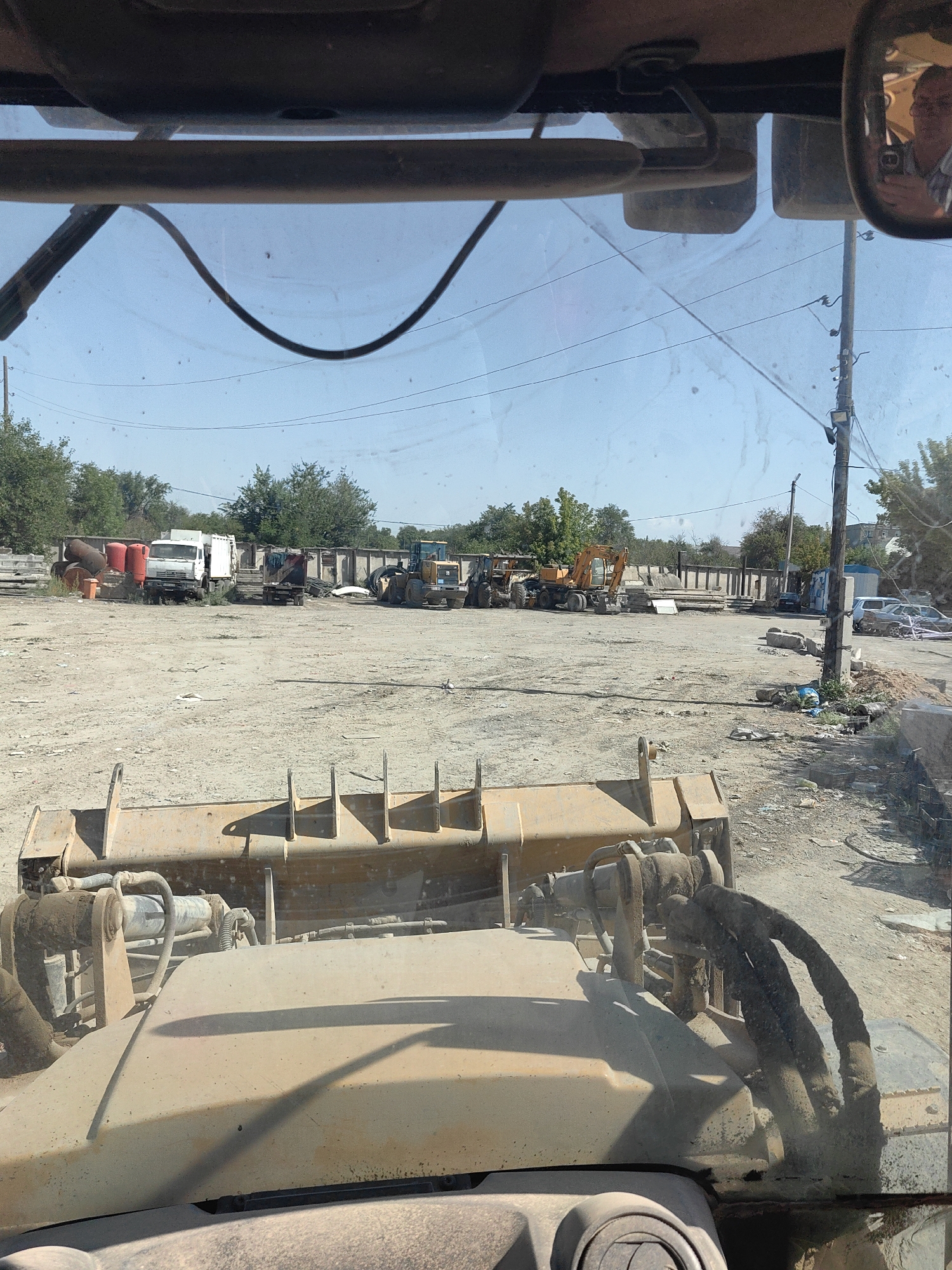 The image shows a view from inside a vehicle, possibly a loader, looking out at a junkyard or construction material storage area in Shymkent, Kazakhstan. The foreground is dominated by the dusty hood and part of the vehicle's attachment. In the middle ground, a variety of heavy machinery is visible, including a yellow front-end loader, a smaller yellow excavator, and what appears to be another excavator or backhoe. There are also several trucks, including a white one. A concrete wall runs along the back of the lot, with trees and other vegetation visible beyond it. Scattered throughout the area are piles of materials, debris, and some cylindrical objects. In the distance, a power pole with wires is present. A side-view mirror on the right shows a reflection of a person holding a phone. The ground is dirt and gravel, with evidence of recent activity.
