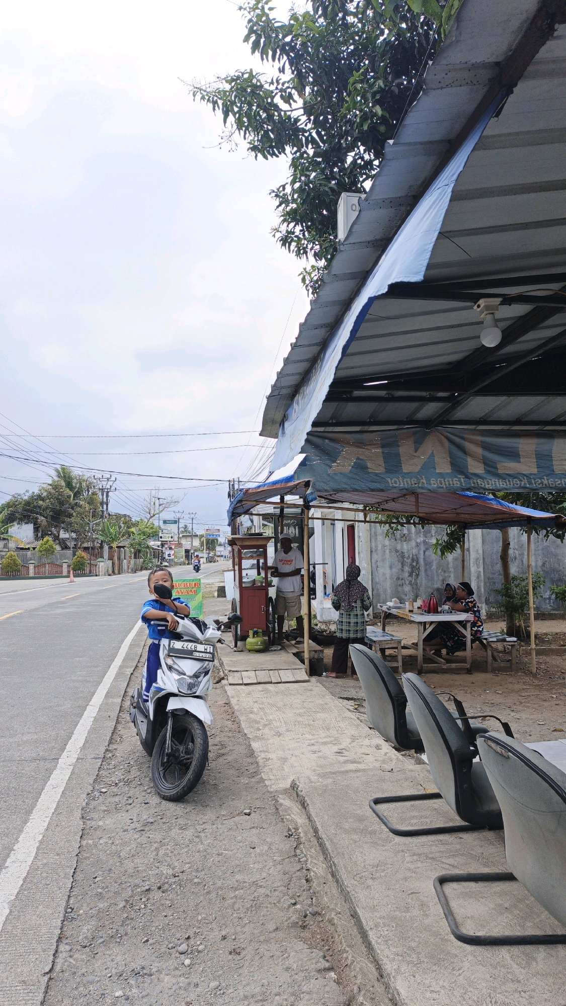 A young boy wearing a blue shirt and a black mask is standing next to a white scooter on the side of a road in Cibenda, Indonesia. He is looking towards the camera. The scooter is parked on a gravel shoulder next to a paved road with a white line. In the background, there is a covered outdoor seating area with tables and chairs, likely a food stall or restaurant. A man in a white t-shirt and shorts is standing behind a food cart, and two women are seated at one of the tables. The area is lined with trees and there are utility poles and power lines visible along the road. The sky is overcast.