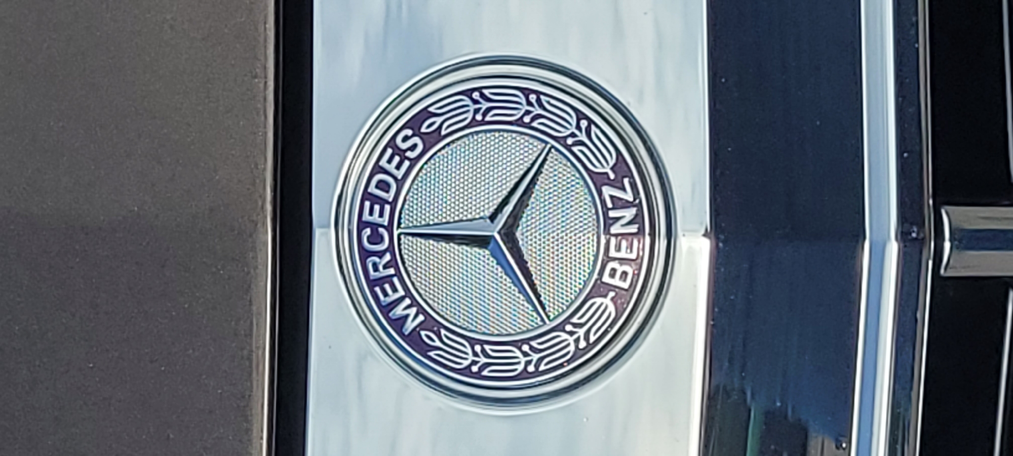 A Mercedes-Benz emblem is prominently displayed on the front of a vehicle, likely a car. The emblem is circular with a silver three-pointed star at its center. The words "MERCEDES" are inscribed in an arc above the star, and "BENZ" is inscribed in an arc below it. The emblem is surrounded by a silver ring with decorative leaf-like elements on either side. The vehicle's dark-colored hood is visible above the emblem, and a chrome grille is visible below. The lighting suggests it is daytime, with reflections of the sky visible on the chrome surfaces. This image does not provide information about a specific location in Riga, Latvia.