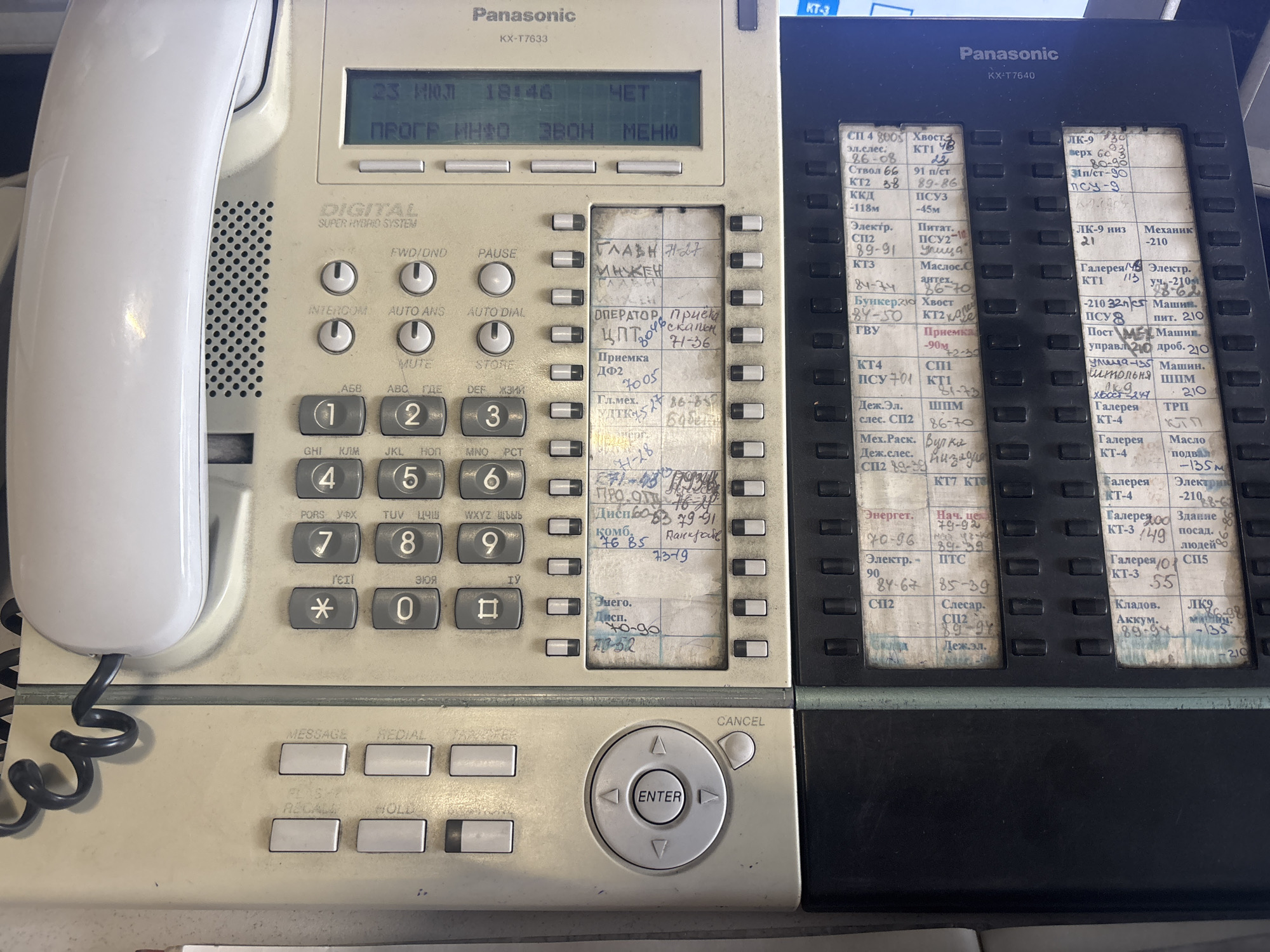 The image shows a close-up, slightly angled view of a beige and black Panasonic KX-T7633 digital super hybrid system telephone. The telephone is situated on a flat, light-colored surface, possibly a desk. The device is in an office or telecommunications environment, as indicated by the telephone itself and the labeling on its keys and display.
The telephone's digital display screen is visible at the top, showing "23 ИЮЛ 18:46 ЧЕТ" on the first line and "ПРОГР ИНФ ЗВОН МЕНЮ" on the second. Below the display are rows of programmable function keys, labeled with various abbreviations and numbers, many of which are handwritten on small paper slips inserted into holders adjacent to the keys. These labels appear to be in Russian and Cyrillic script.
The left side of the telephone features a prominent white handset. The keypad section of the telephone includes numbered keys from 0 to 9, as well as asterisk and hash keys. Below the keypad are function buttons such as "MESSAGE," "REDIAL," "HOLD," and "RECALL." A circular navigation control with an "ENTER" button is positioned in the lower right portion of the main telephone unit.
The right side of the telephone features a separate section with more programmable function keys and corresponding handwritten labels. These labels also appear to be in Russian and Cyrillic script, indicating various departments or functions, such as "Эл.лес," "КП," "КТ2," "КТЗ," "Энерг," "Сл/С," and numerical codes.
The overall condition of the telephone suggests it is a well-used office device. The lighting in the scene is consistent with indoor office lighting. The image does not provide any explicit information about the location being the city of Kryvyi Rih, Ukraine, or any specific details about people, actions, or interactions occurring at the time the photograph was taken. The context is purely based on the object itself and its typical use.