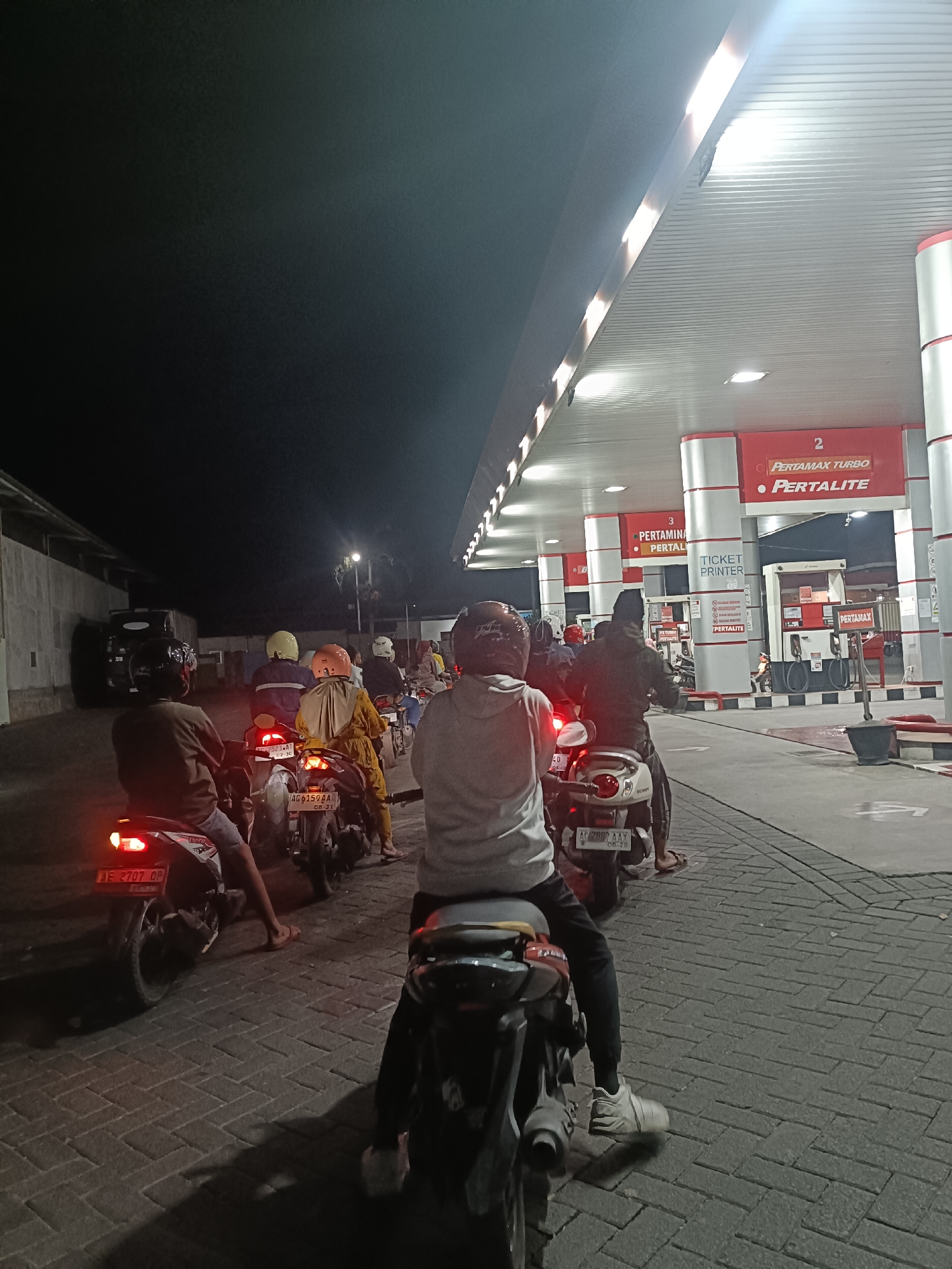 The image depicts a nighttime scene at a Pertamina gas station, likely located in Kediri City, Indonesia. A queue of motorcycles with riders is waiting to refuel. The gas station is illuminated by overhead lights, highlighting the red and white branding of Pertamina. Several motorcycles are visible, with their license plates showing Indonesian characters. People are seated on their motorcycles, some wearing helmets. The ground is paved with interlocking bricks. The overall atmosphere suggests a routine evening at a public service facility.
