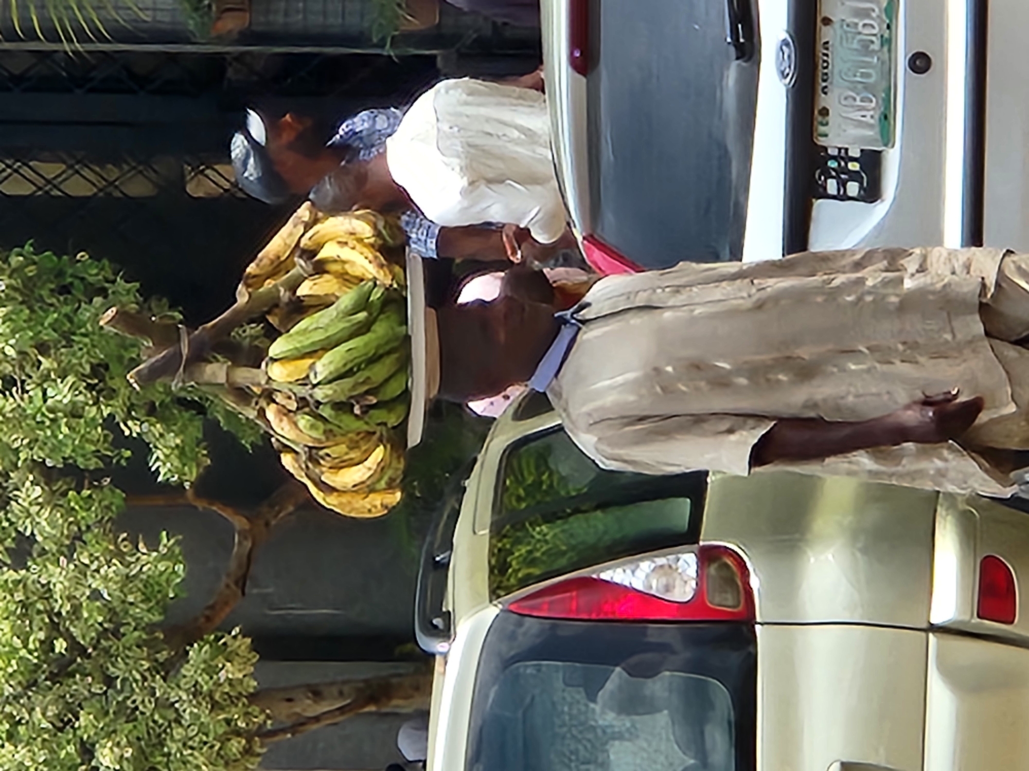 The image captures a street scene in Jos, Nigeria, likely near a market or thoroughfare, given the presence of vendors and vehicles. In the foreground, a man wearing a light brown, loosely fitting garment with a blue collar is seen from the waist up, facing left, and appears to be looking downwards. He has bananas balanced on his head, supported by a flat, circular object. Behind him, slightly to the right, another man is visible, wearing a white shirt and a dark baseball cap, looking to his right. His back is mostly to the camera. Two cars are present; a light-colored SUV is on the left, and a silver car is on the right, displaying a license plate with the characters "YAB 615BJ" and the text "FED. ABUJA". The background features a tree with green and light-colored leaves, suggesting a sunny day. The overall atmosphere appears to be one of everyday commerce and urban life.